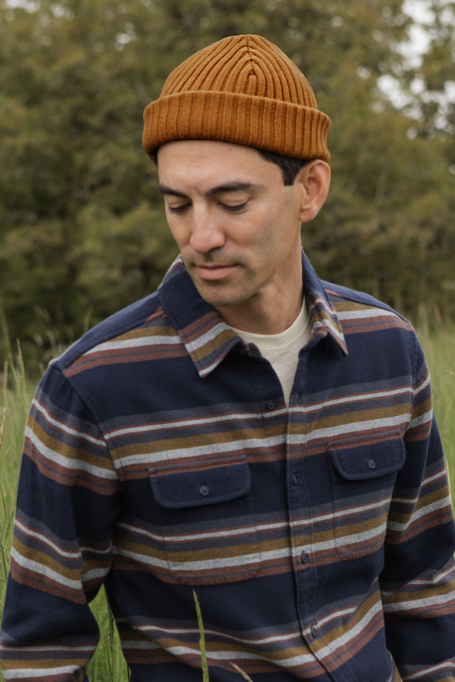 A man wears the Bridge & Burn Chunky Ribbed Beanie in Warm Ochre and a blue striped flannel shirt, standing outdoors in tall grass with trees blurred in the background, looking down with a neutral expression.