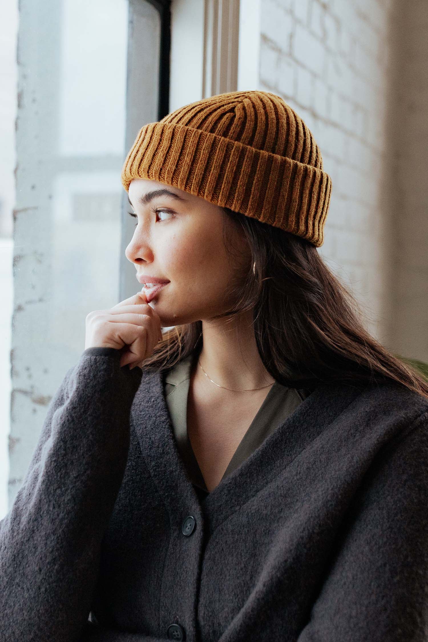A woman in a Bridge & Burn Chunky Ribbed Beanie in Warm Ochre and a dark coat sits by a window, gazing outside thoughtfully with her hand near her mouth.