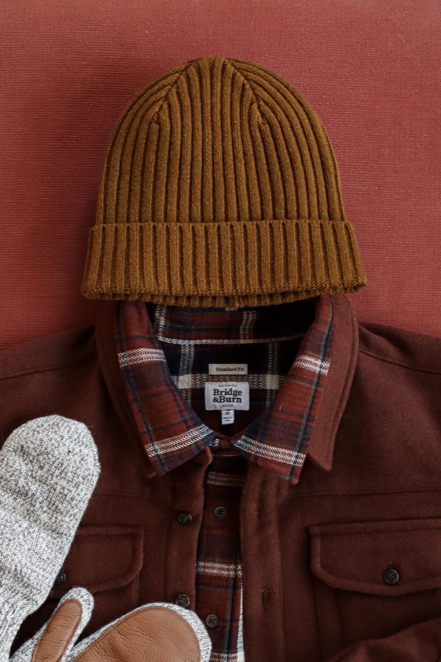 A Bridge & Burn Chunky Ribbed Beanie in Warm Ochre, a brown flannel shirt with a plaid collar, and gray and brown mittens are arranged flat on a reddish surface.