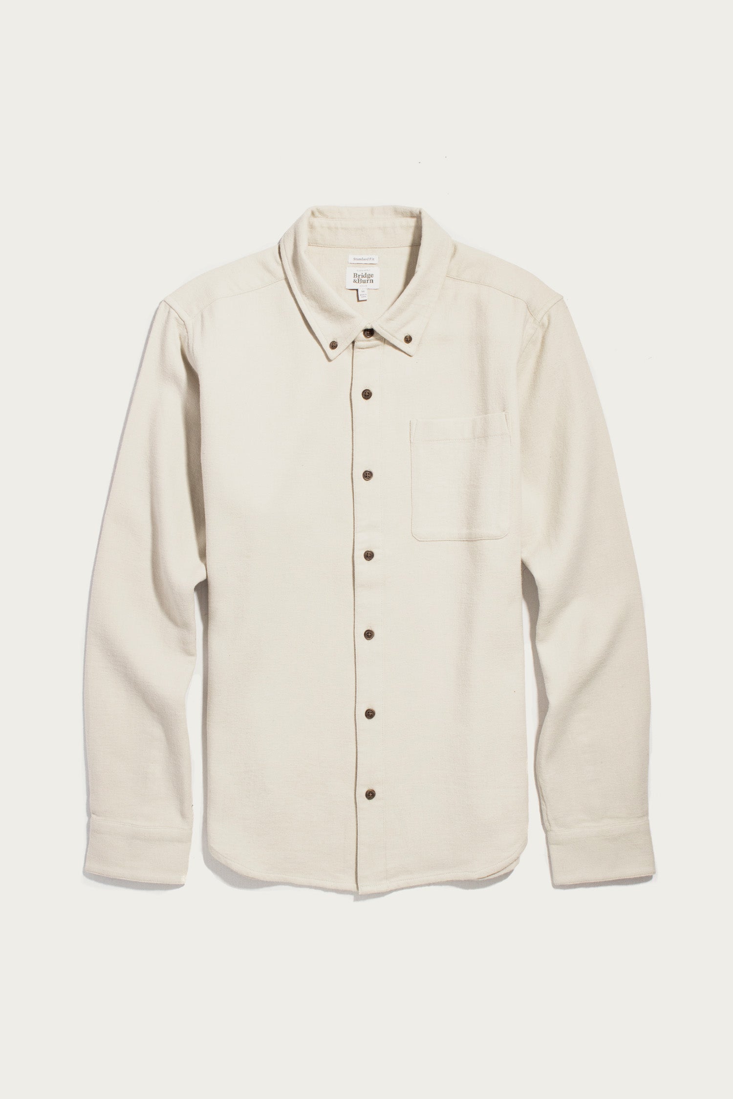 The Bridge & Burn Sutton Slim Shirt in Fawn Flannel is a slim-fit, long-sleeve shirt crafted from cream-colored organic cotton with a soft flannel texture. It features a single chest pocket, dark buttons, and is showcased on a plain white background for a clean, minimalist look.