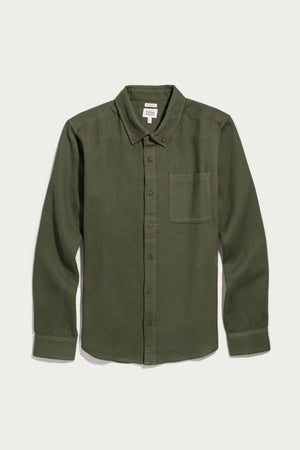 Bridge & Burn’s Sutton Slim Shirt in Pine Flannel features an olive green button-up design with long sleeves, a chest pocket, button-down collar, and brown buttons. Laid flat on a white background, this minimalist organic cotton shirt offers timeless style and comfort.