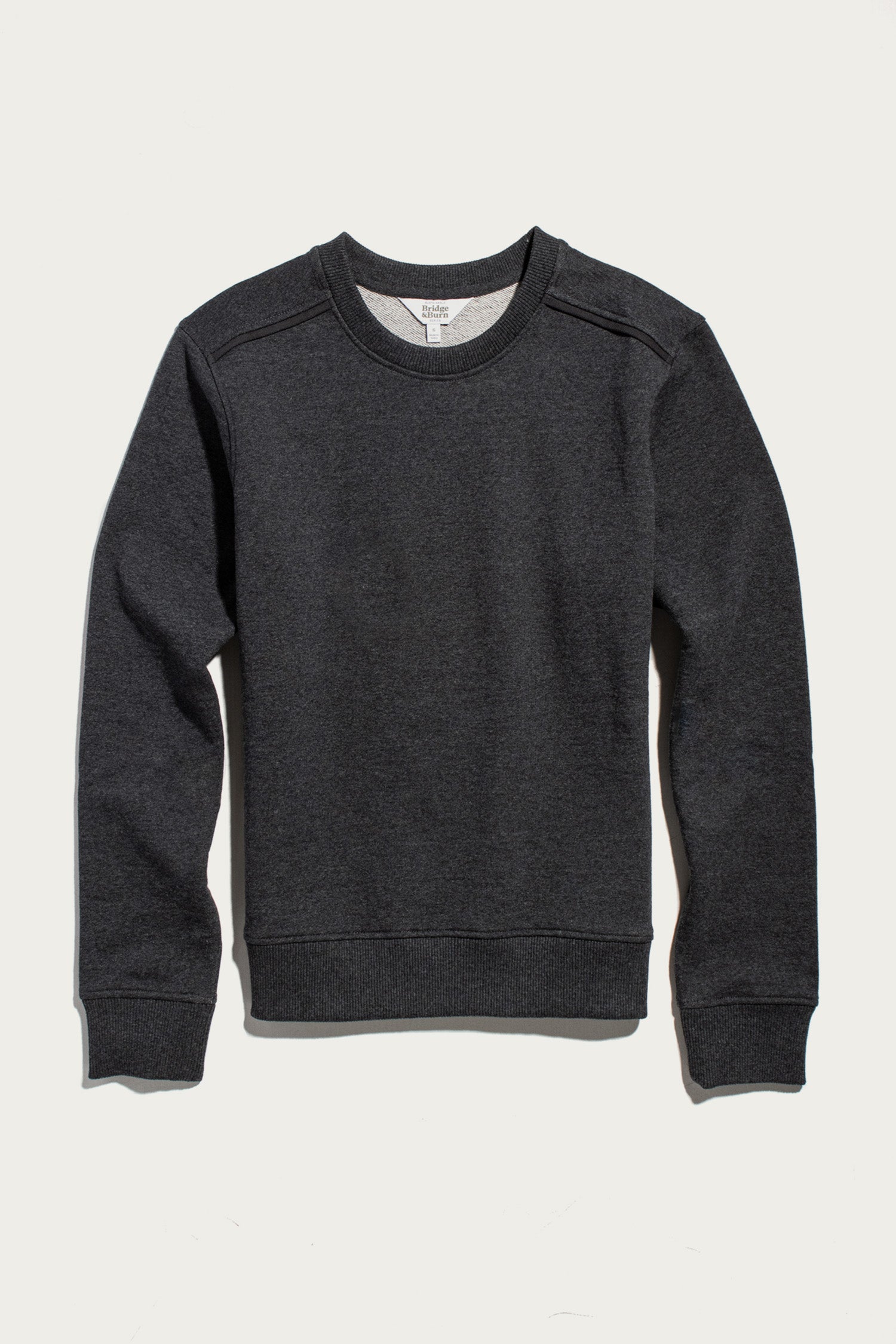 The Bridge & Burn Bailey Crew Sweatshirt in Graphite, a sustainable long-sleeve crewneck with ribbed cuffs and hem, is laid flat on a light background. Simple seams run along the sleeves and shoulders.