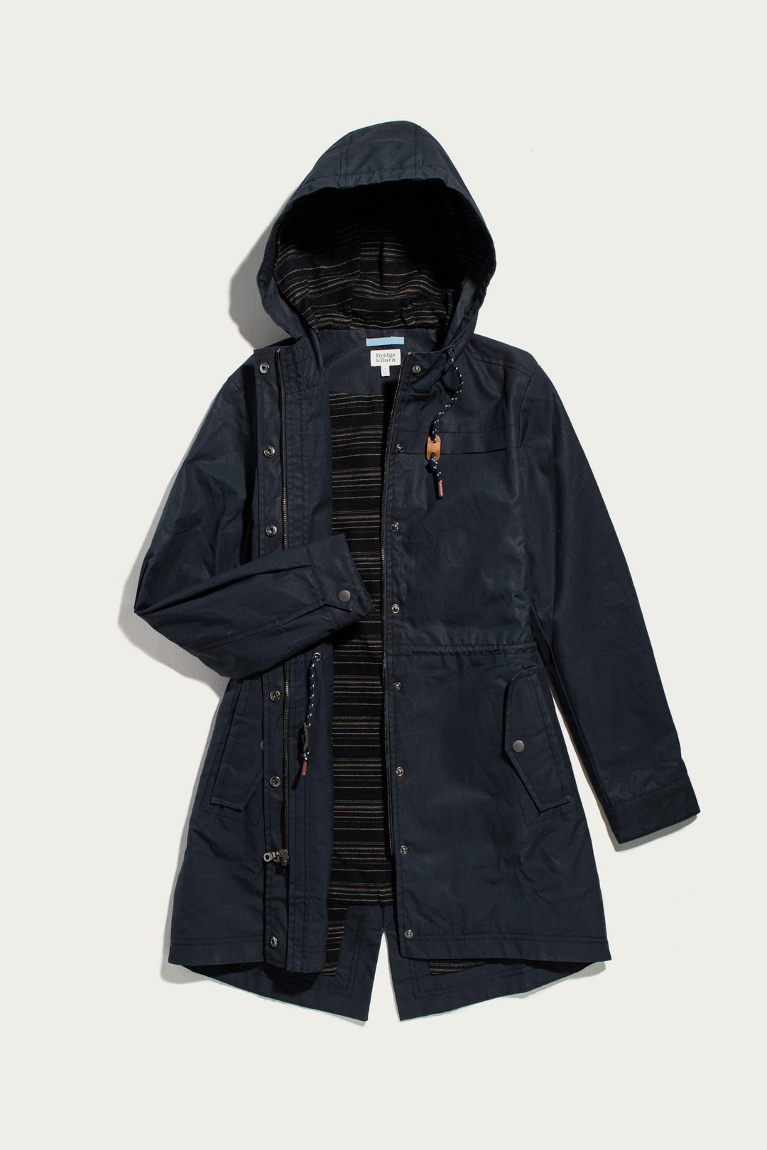 Bridge & Burn Briar Jacket in navy waxed cotton featuring a hooded parka silhouette, striped interior lining, drawstring waist, and front zipper with snap closures.