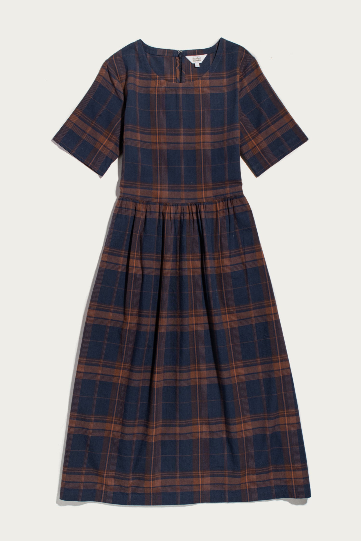 Introducing the Perry Dress in Cinder Plaid from Bridge & Burn—a midi-length dress crafted from organic cotton, featuring short sleeves, a round neckline, and a navy blue and brown plaid pattern. The gathered waist creates a subtly flared skirt for an effortlessly flattering silhouette.