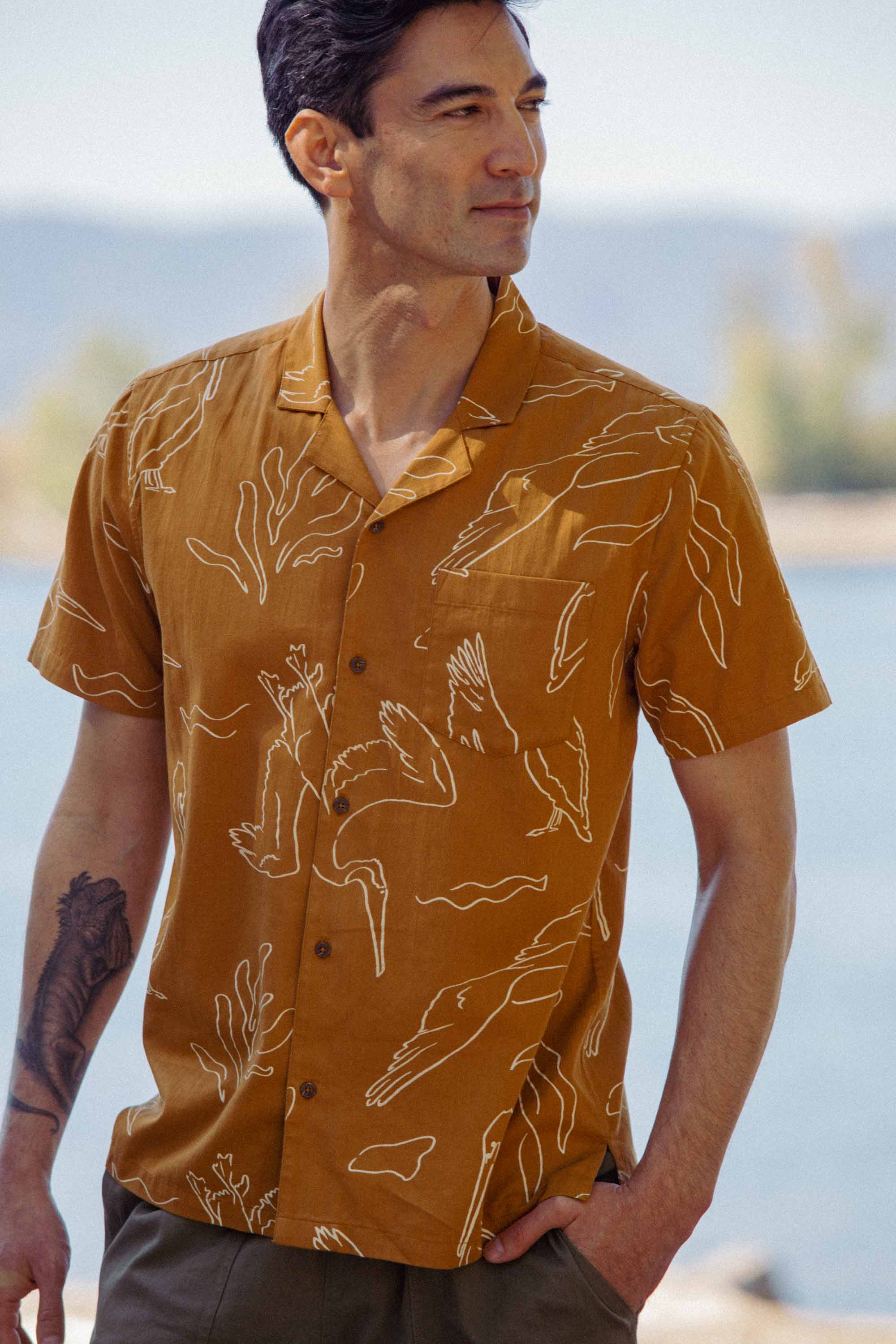 A man stands outdoors by a body of water, looking to the side, wearing the Bridge & Burn Bodie Shirt in Shoreflight—a short-sleeved retro camp collar shirt with a brown-and-white abstract line pattern. Trees and mountains blur in the background.