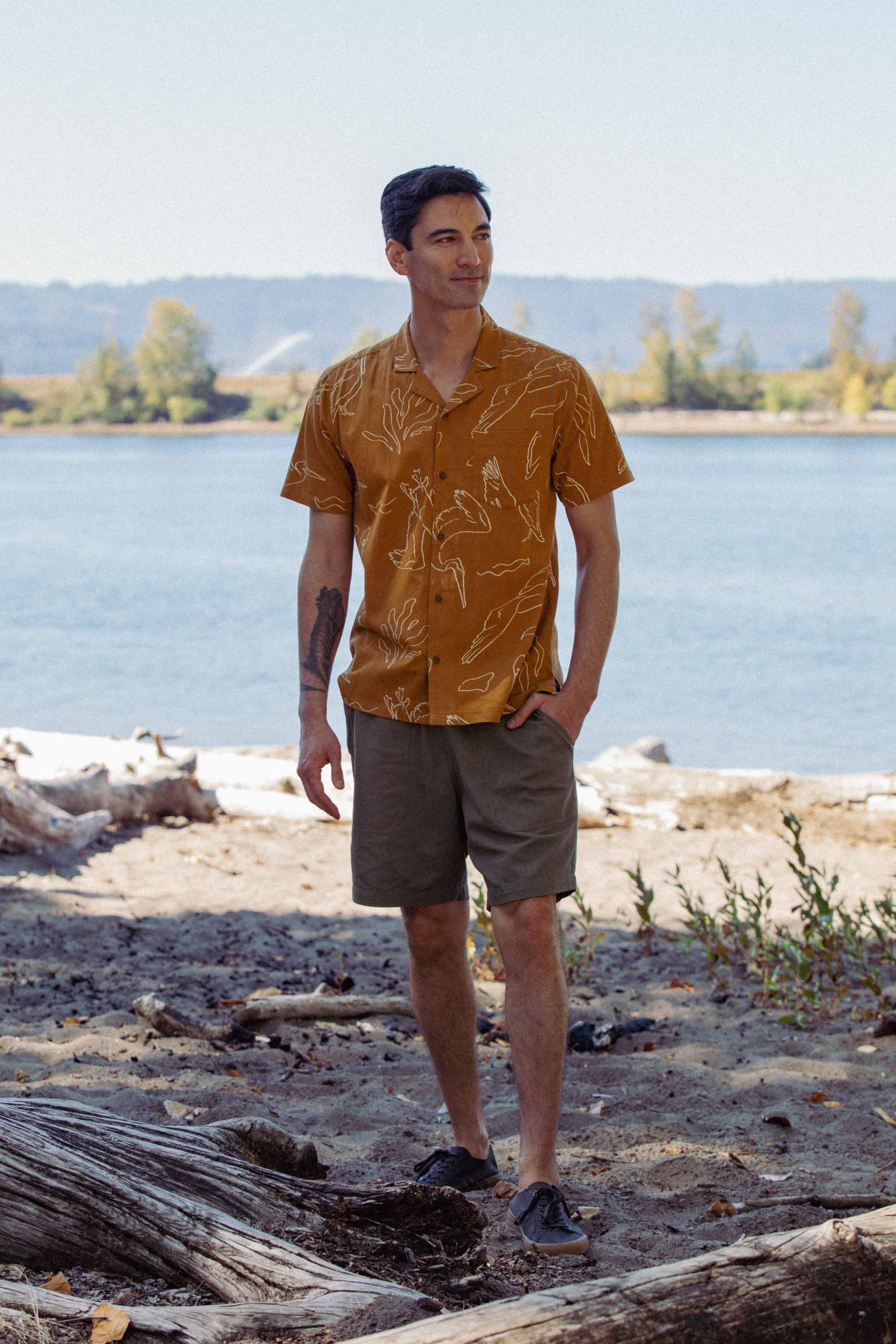 A man stands on a sandy riverbank with trees behind him, wearing the Bridge & Burn Bodie Shirt in Shoreflight, olive shorts, and dark shoes. One hand is in his pocket as he gazes off to the side.