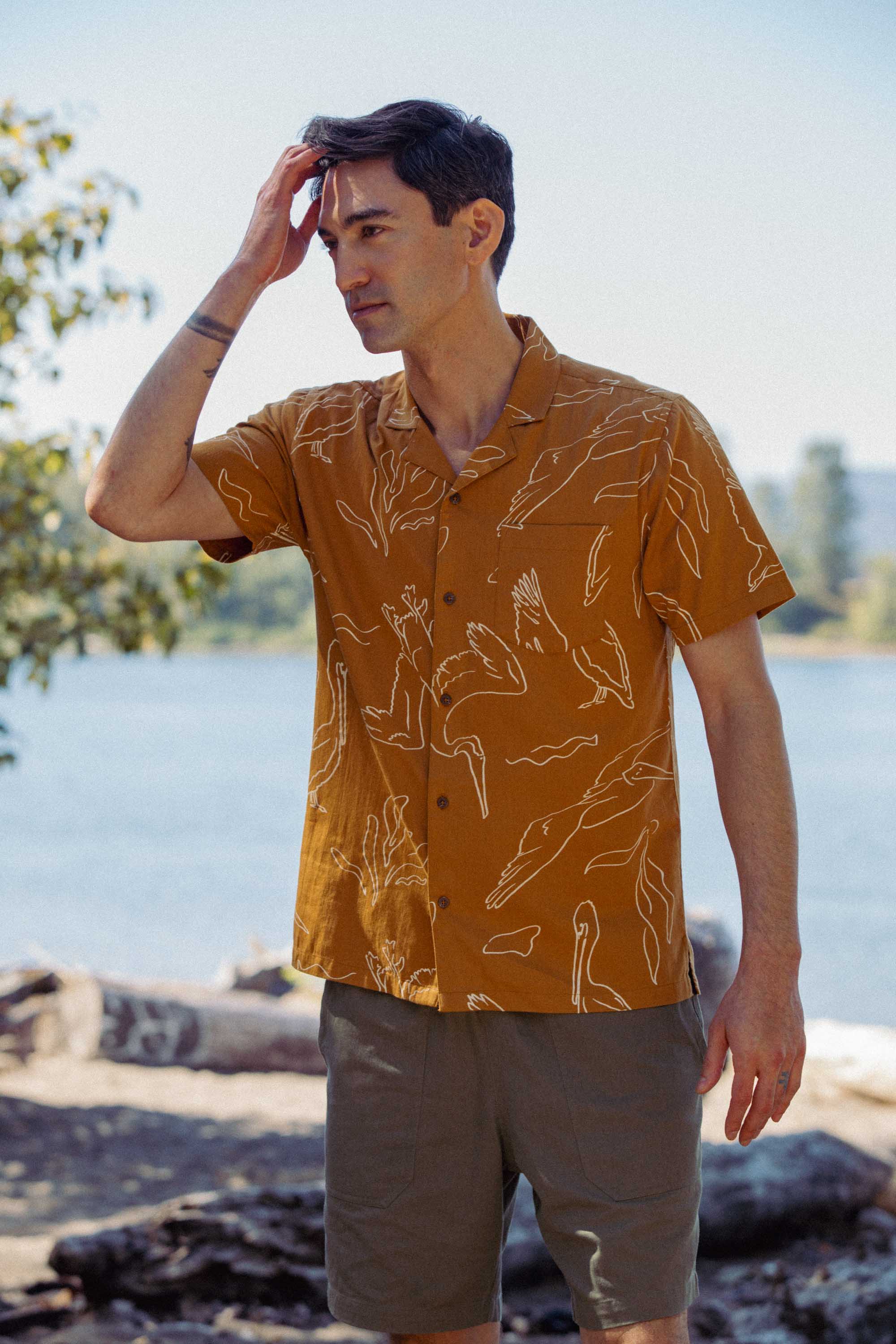 A man stands outdoors by the water in a Bridge & Burn Bodie Shirt / Shoreflight, featuring mustard fabric with white animal line drawings, paired with gray shorts. He touches his hair as trees and a sunny sky fill the background.