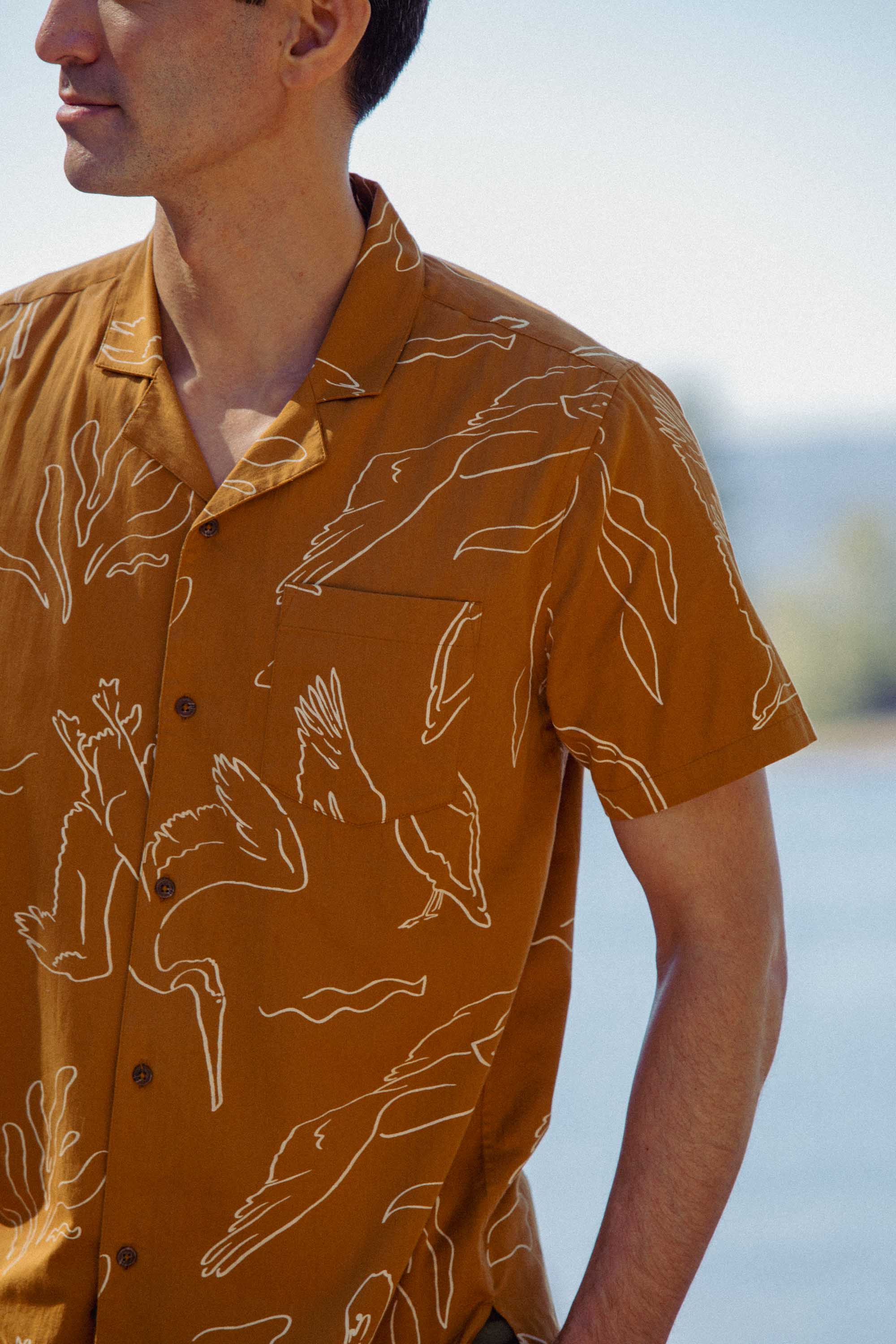 A man wears the Bridge & Burn Bodie Shirt in Shoreflight, a brown lightweight summer camp collar shirt featuring white abstract bird line drawings. He stands outdoors near water with only his upper body visible, background blurred.