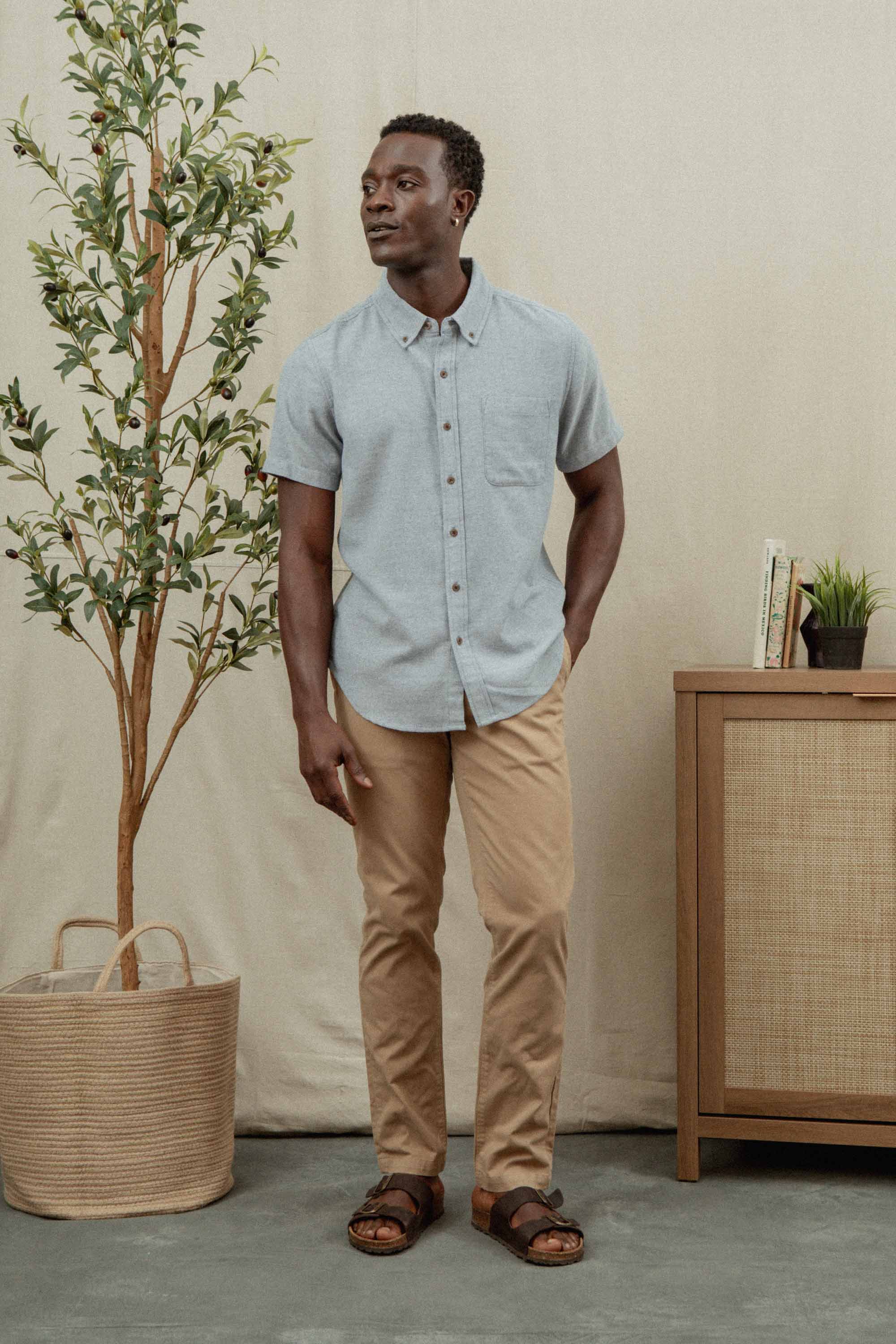 A man wearing the Bridge & Burn Bryce Shirt in Denim Twill stands indoors by a potted plant and cabinet, displaying a stylish, sustainable look.