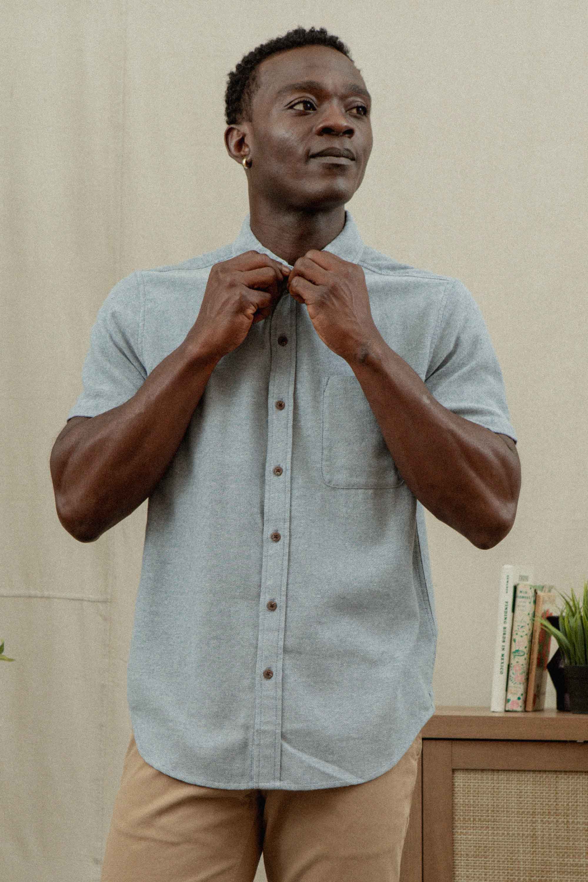 A man indoors buttons the collar of his Bridge & Burn Bryce Shirt in Denim Twill. He wears tan pants with a neutral expression, while a cabinet and books in the background hint at his thoughtful, sustainable wardrobe choices.