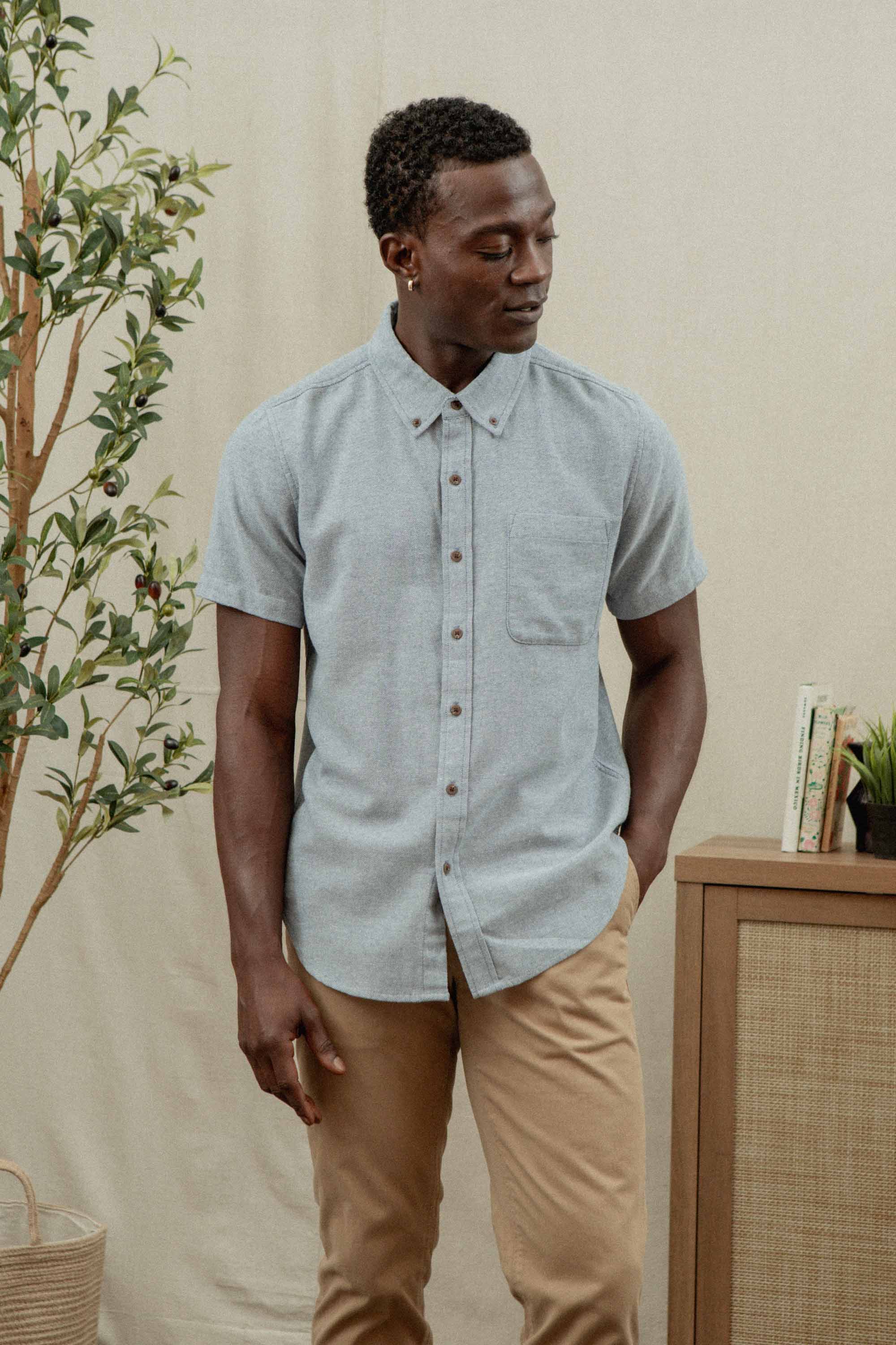 A man stands indoors by a leafy plant and wooden cabinet, wearing Bridge & Burn’s Bryce Shirt in Denim Twill and khaki pants, showcasing a sustainable wardrobe.