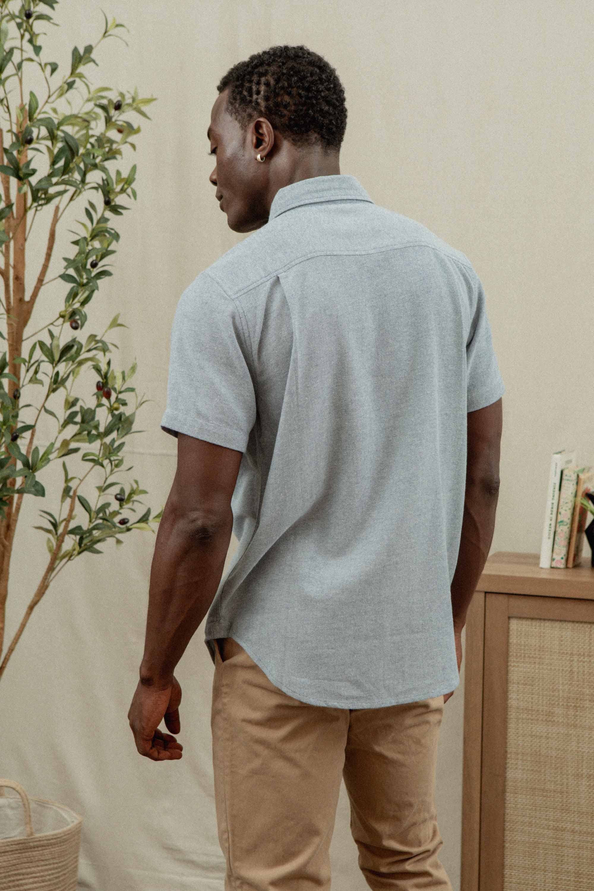 A man stands with his back to the camera in a neutral-toned room, wearing the Bryce Shirt in Denim Twill by Bridge & Burn and beige pants, creating a sustainable wardrobe vibe alongside a plant and books on a cabinet.
