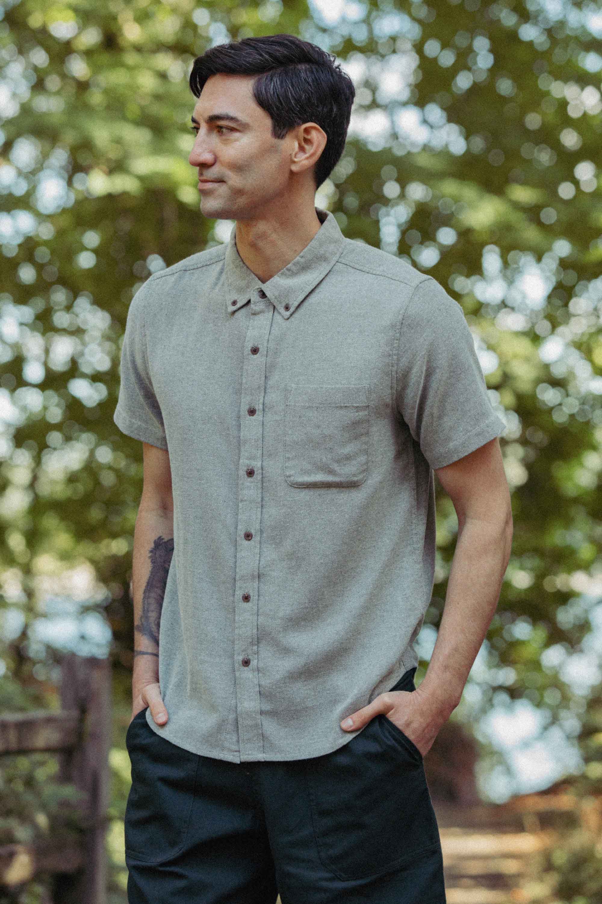 A man with dark hair stands outdoors in the Bryce Shirt in Olive Twill by Bridge & Burn—a short-sleeve button-up with a chest pocket—paired with dark pants. Hands in pockets, he looks to the side amid sunlit trees.