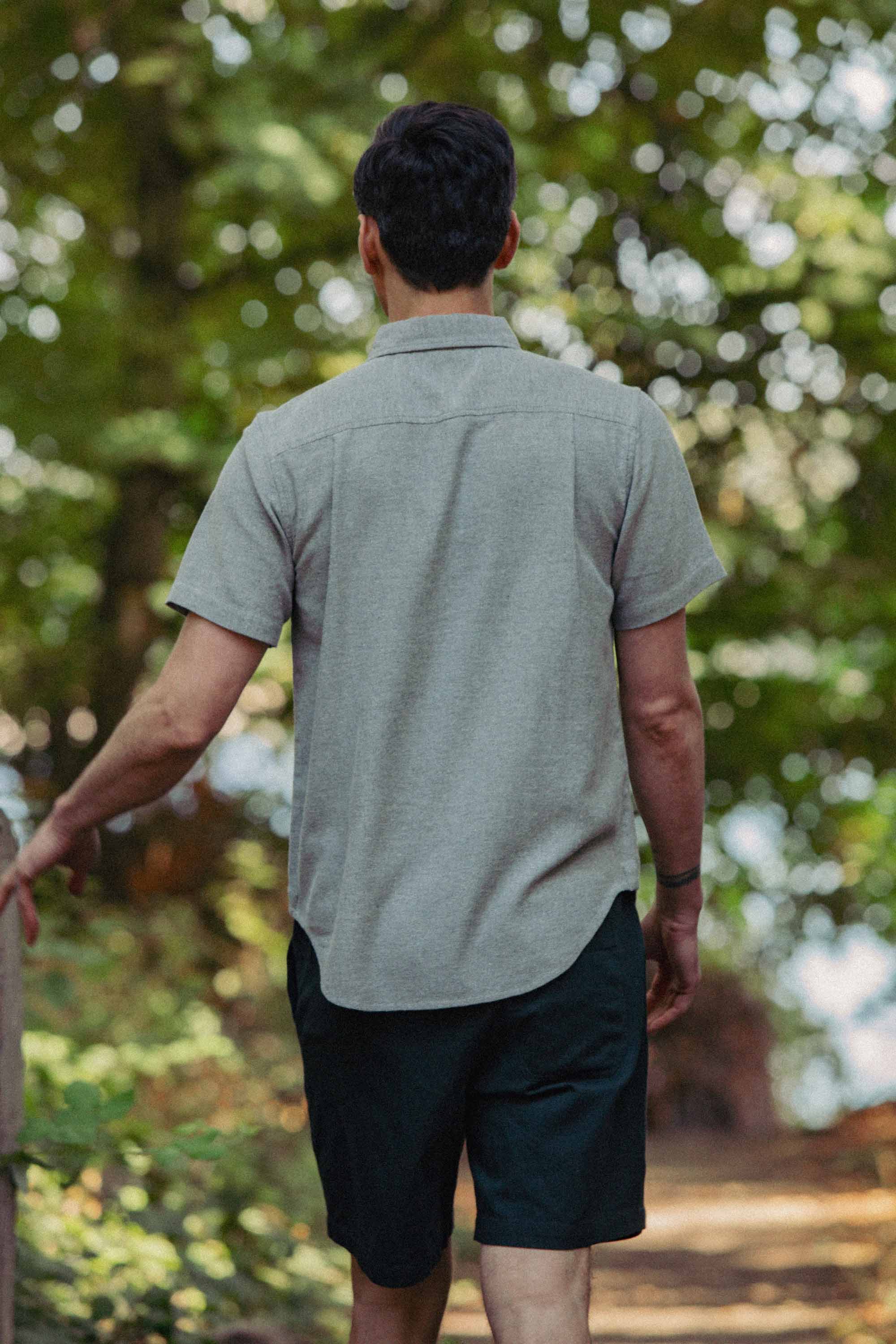A man with short dark hair, wearing the Bridge & Burn Bryce Shirt in Olive Twill and dark shorts, walks away on a forest path surrounded by green trees.