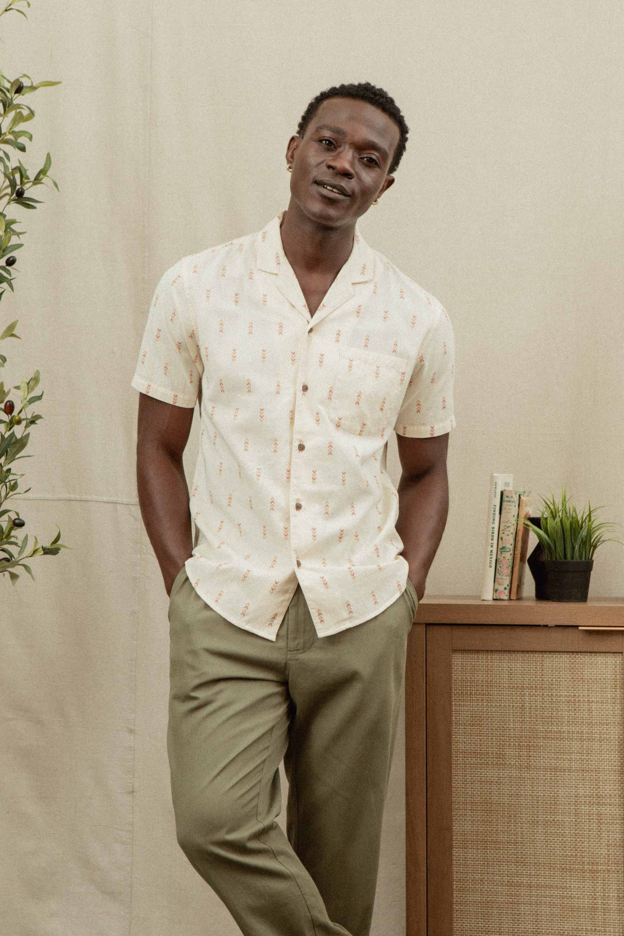 A man stands casually with hands in pockets, wearing the Bridge & Burn Collins Slim Shirt in Dune Chevron and olive green pants, in front of a cabinet with books, a plant, and a neutral backdrop.