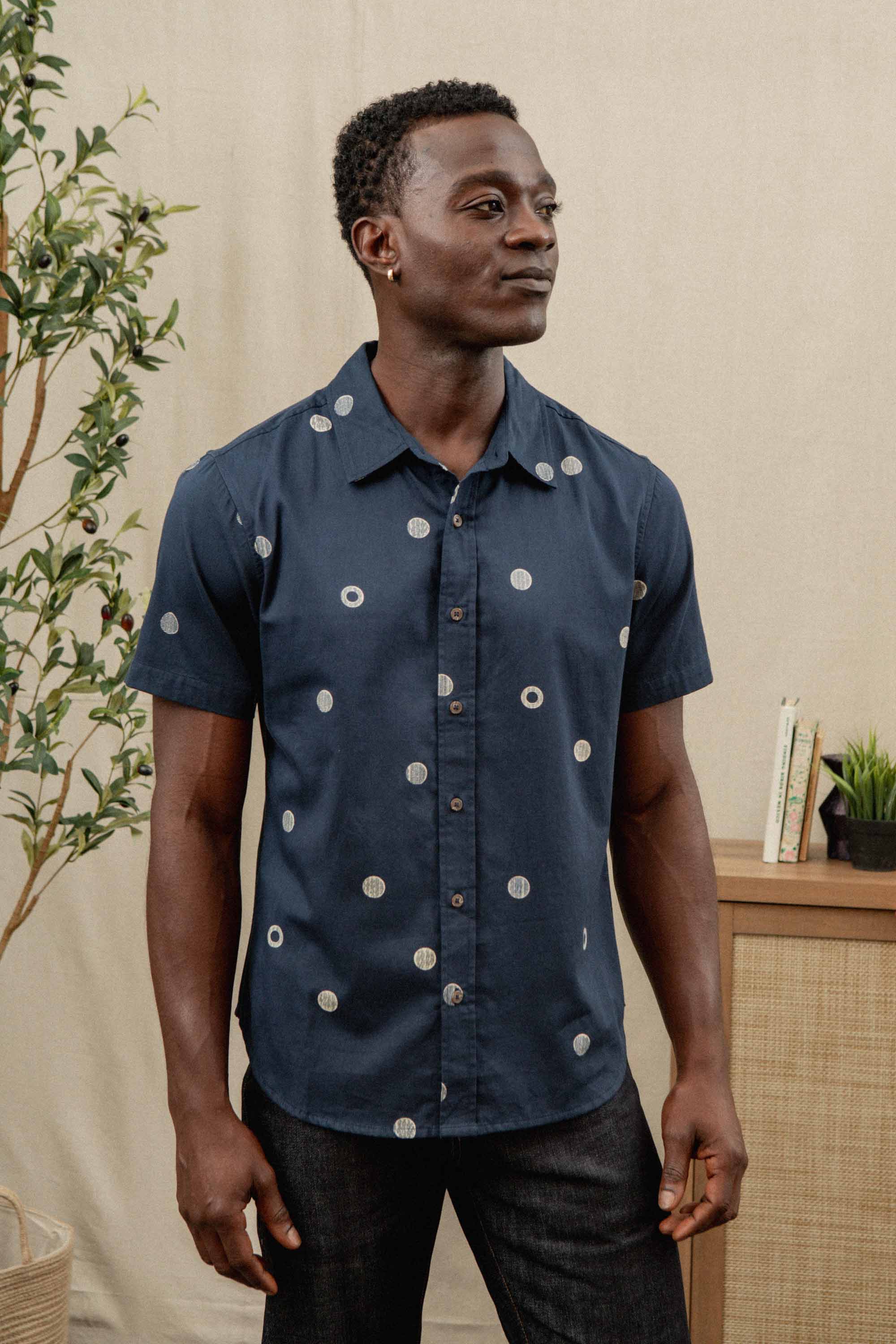 A man in the Bridge & Burn Emerson Slim Shirt / Lunar Polka stands indoors by a plant and a wooden cabinet topped with books and a small potted plant.