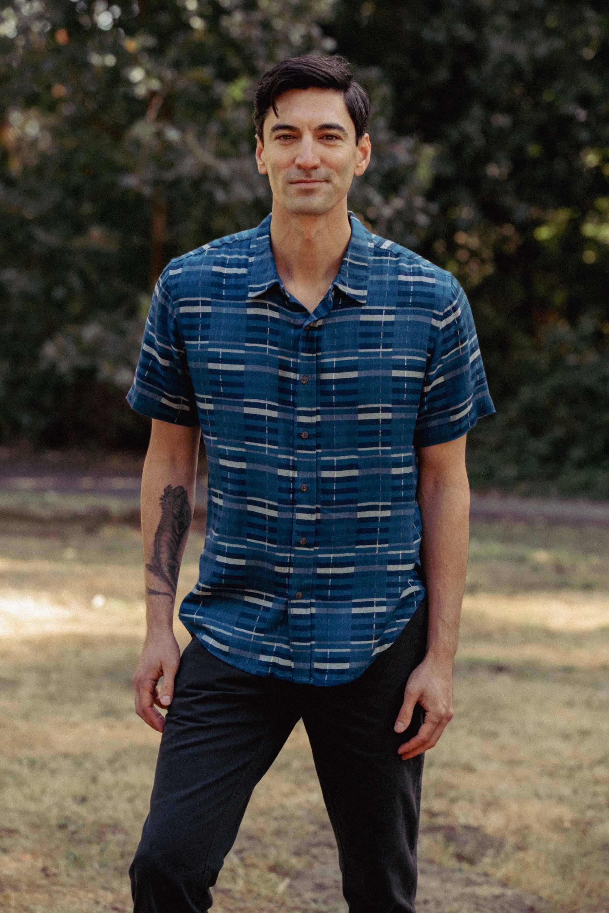 A man with short dark hair smiles outdoors on grass, wearing the Bridge & Burn Emerson Slim Shirt in Stormcrest Stripe and dark pants. He has a tattoo on his left forearm, with trees and greenery in the background.