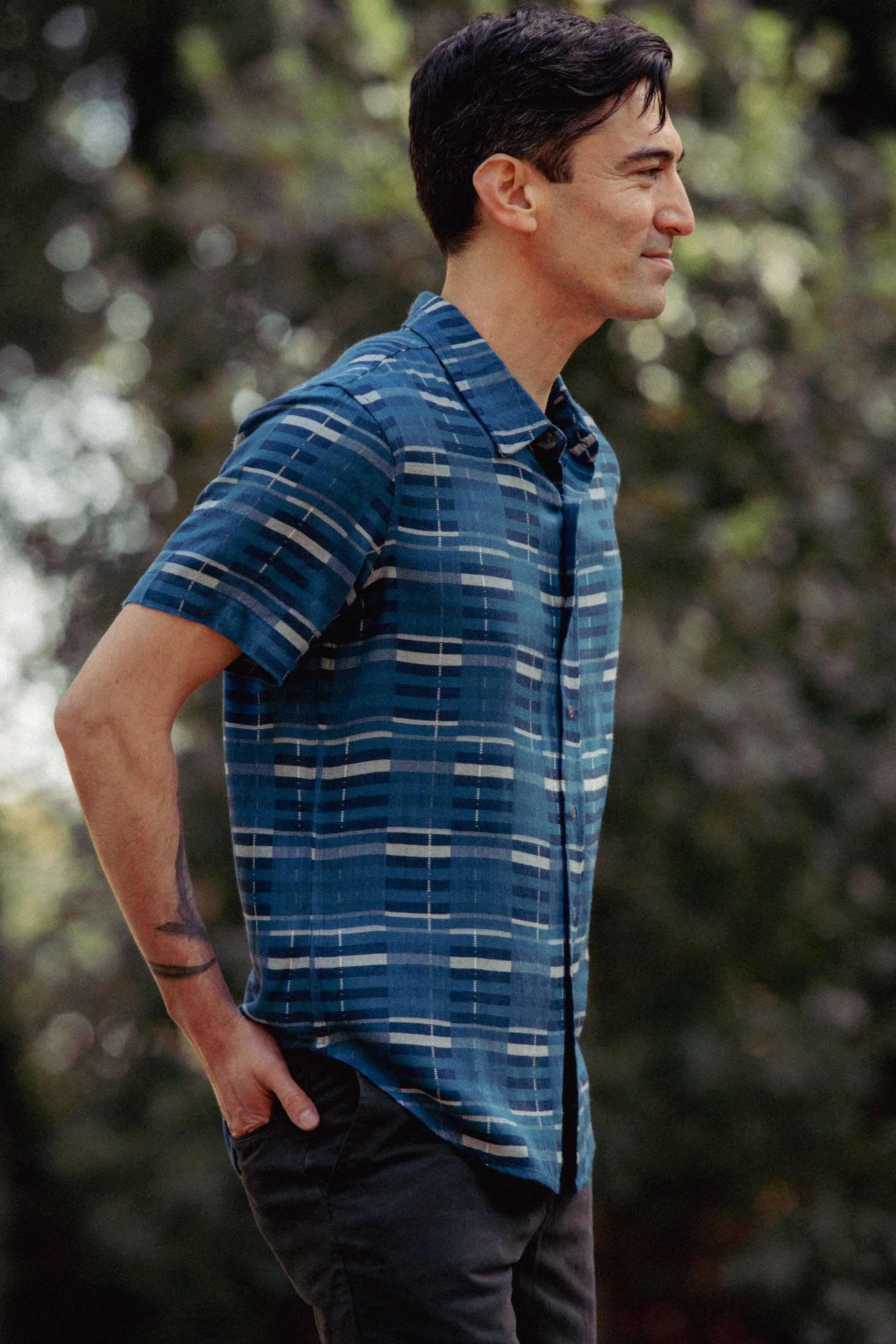 A man with short dark hair stands outdoors, wearing the Bridge & Burn Emerson Slim Shirt in Stormcrest Stripe and dark pants, his right hand in his pocket. Blurred trees and greenery appear in the background.
