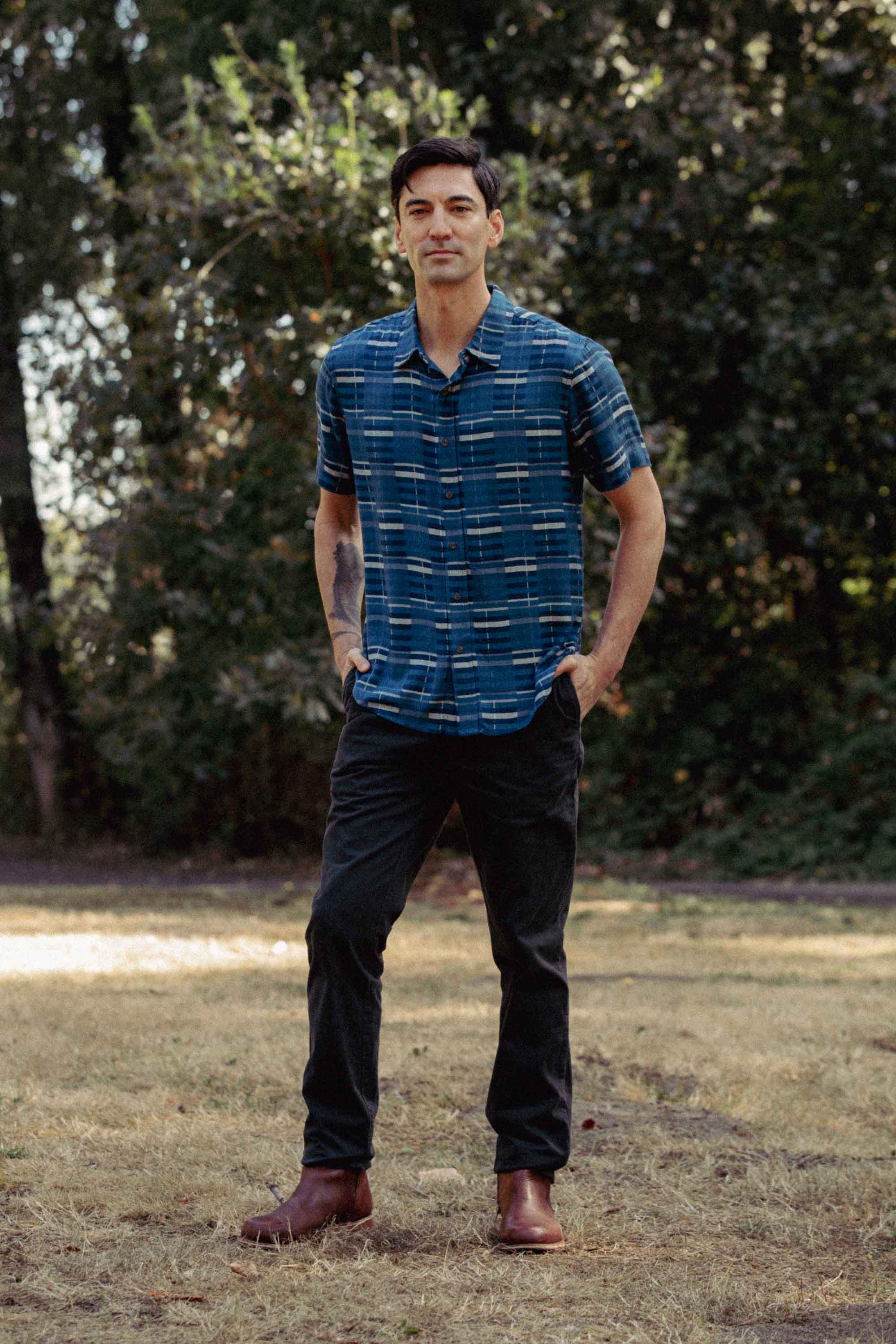 A man stands on dry grass with trees behind him, wearing the Bridge & Burn Emerson Slim Shirt in Stormcrest Stripe, dark pants, and brown boots. He faces the camera, hands in pockets, with a slight smile.