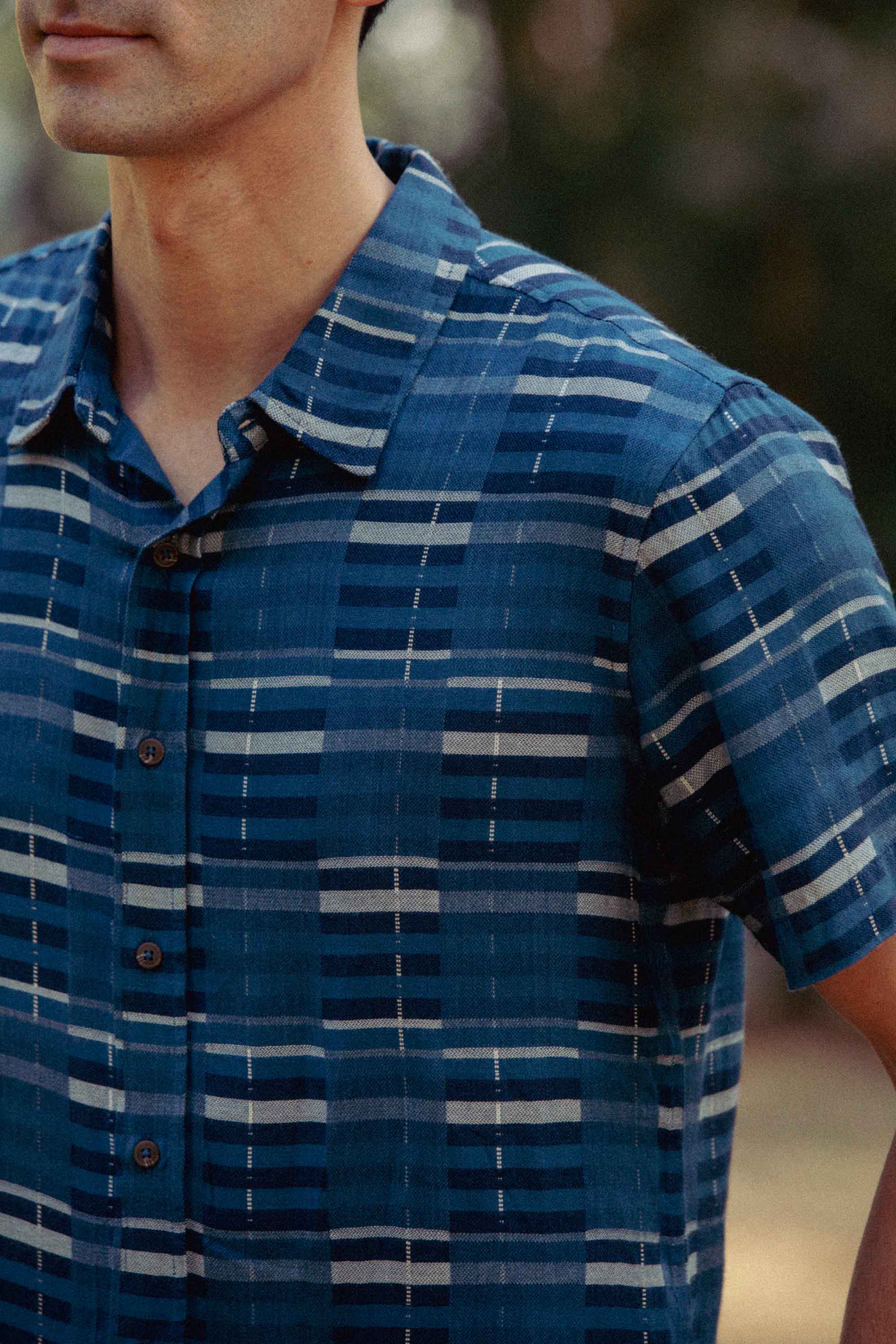 A person is seen outdoors wearing the Bridge & Burn Emerson Slim Shirt in Stormcrest Stripe, a short-sleeve, button-up slim fit style. Only their upper torso and part of the face are visible.