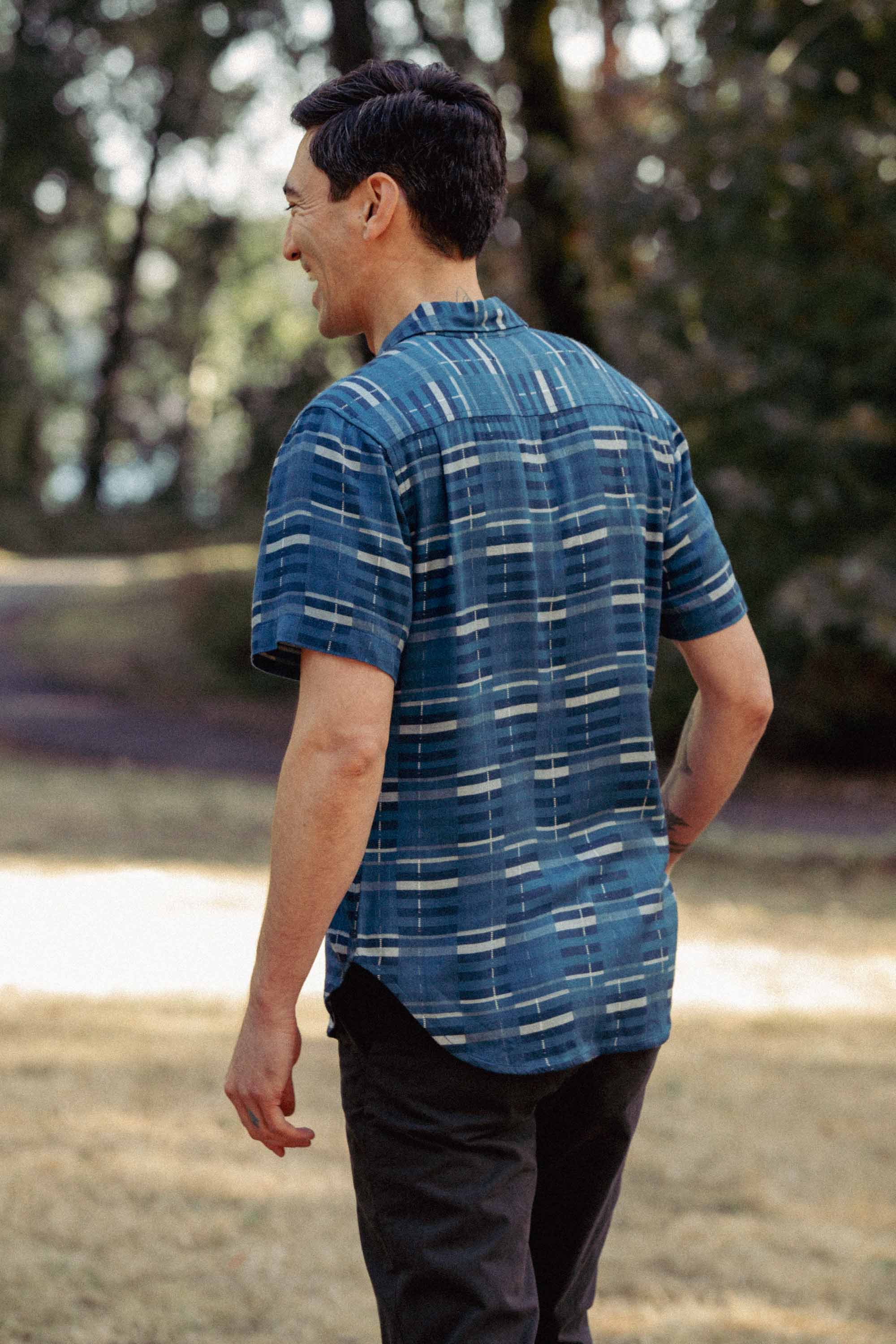 A man stands on grass, smiling with his back turned, wearing Bridge & Burn’s Emerson Slim Shirt in Stormcrest Stripe and dark pants. Trees and a path are softly blurred in the background.
