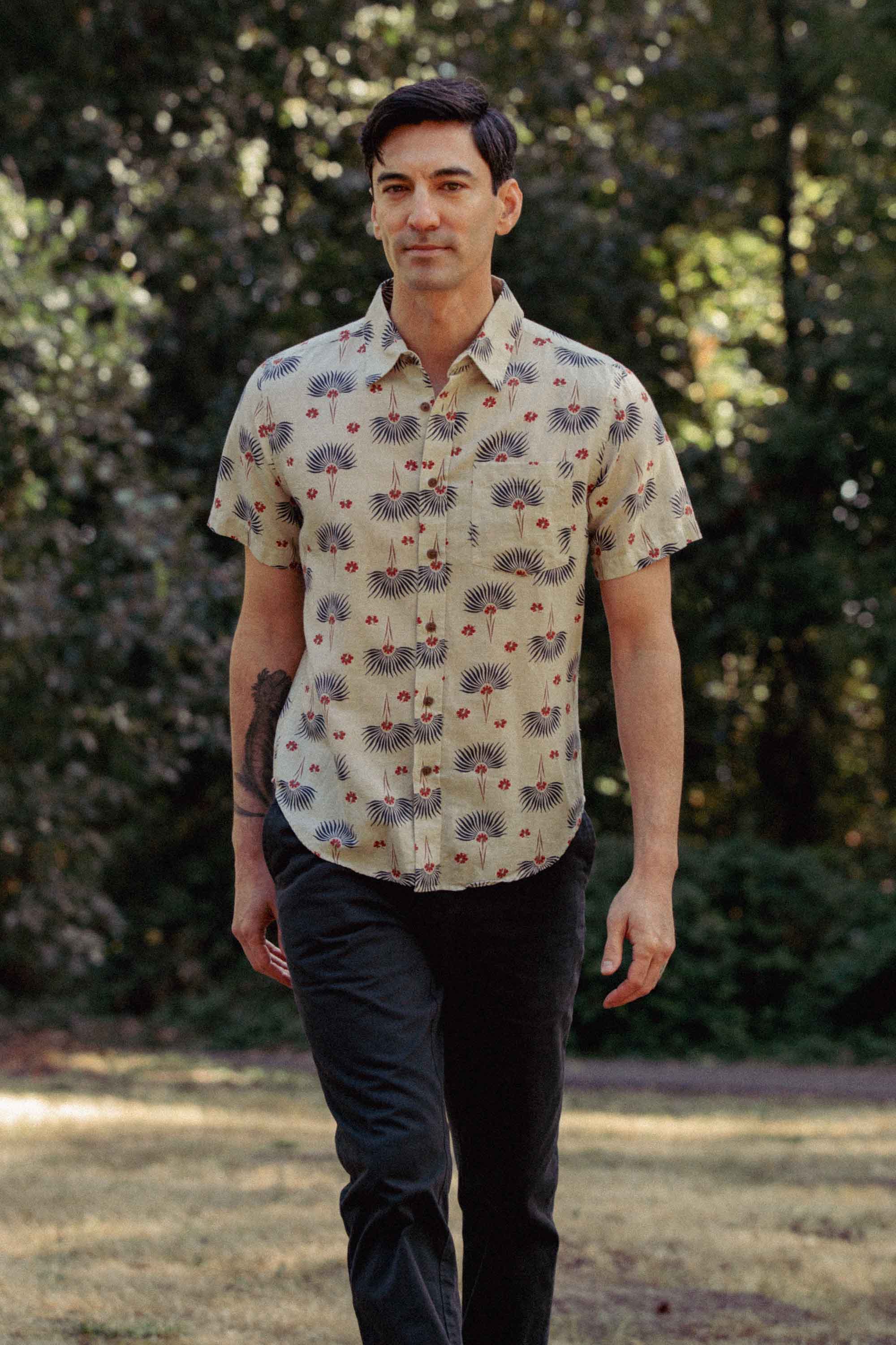 A man with dark hair walks on grass among trees and sunlight, wearing the Bridge & Burn Harbor Slim Shirt in Aloha Floral and dark pants.