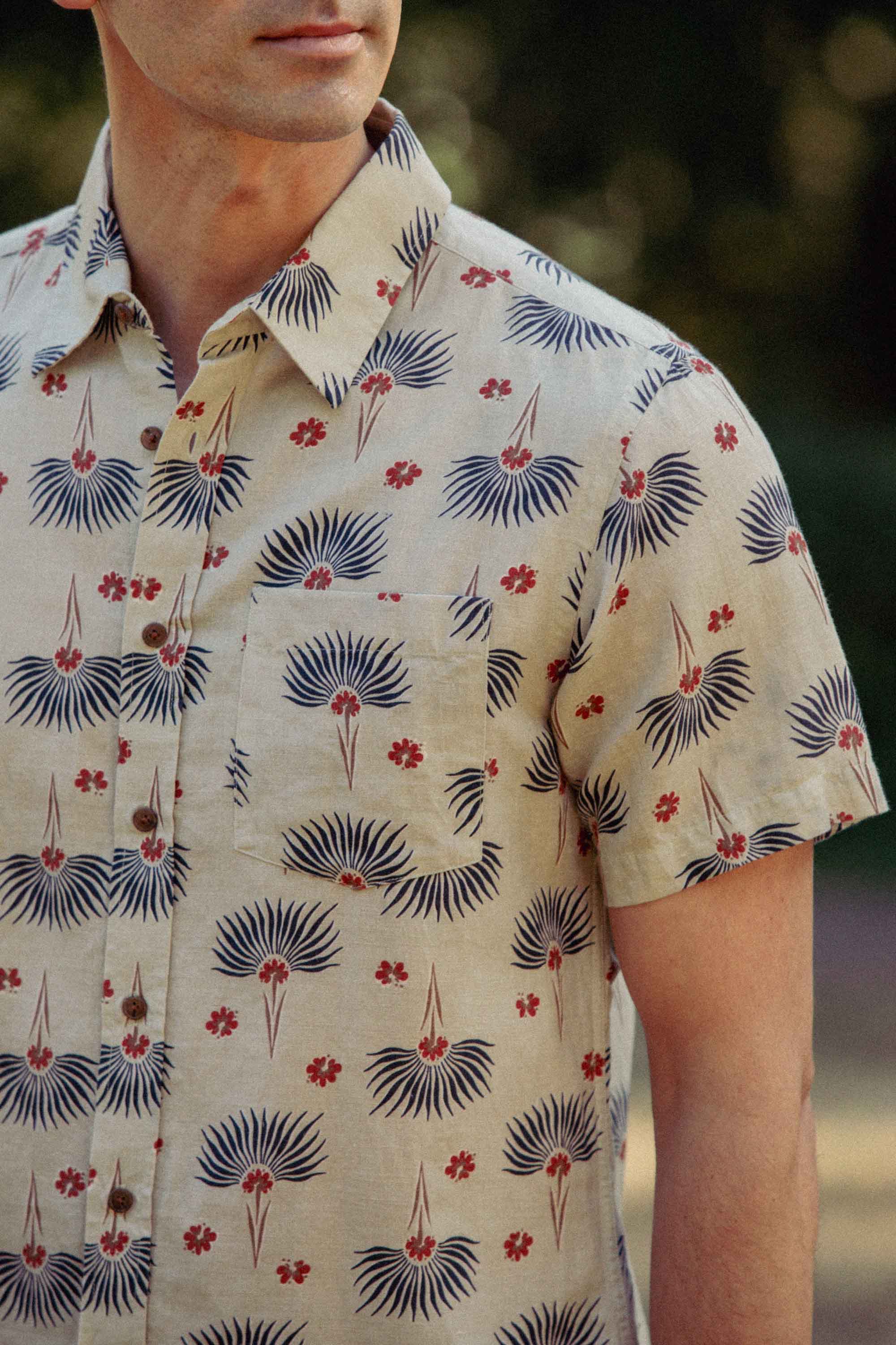 Wearing the Bridge & Burn Harbor Slim Shirt in Aloha Floral, featuring blue fan motifs and red flowers on light fabric, a person stands outdoors with only their upper body and lower face visible.