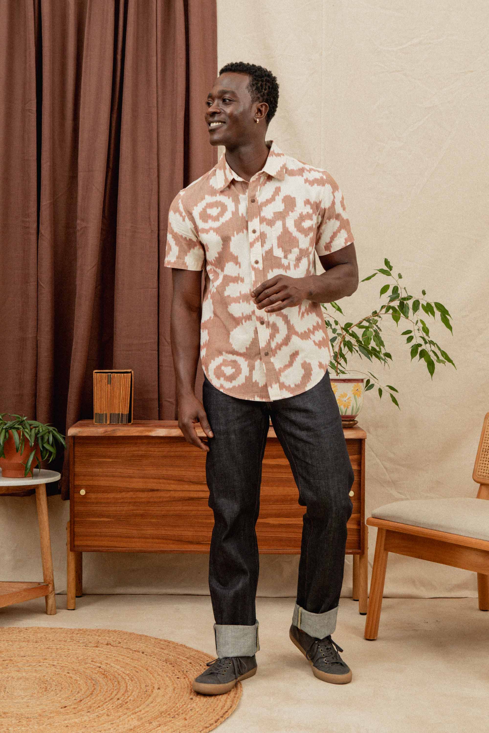A man stands indoors smiling and looking to the side, wearing the Bridge & Burn Harbor Slim Shirt in Pop Leaf and dark cuffed jeans. He is next to a mid-century wooden console, a potted plant, and a brown curtain.