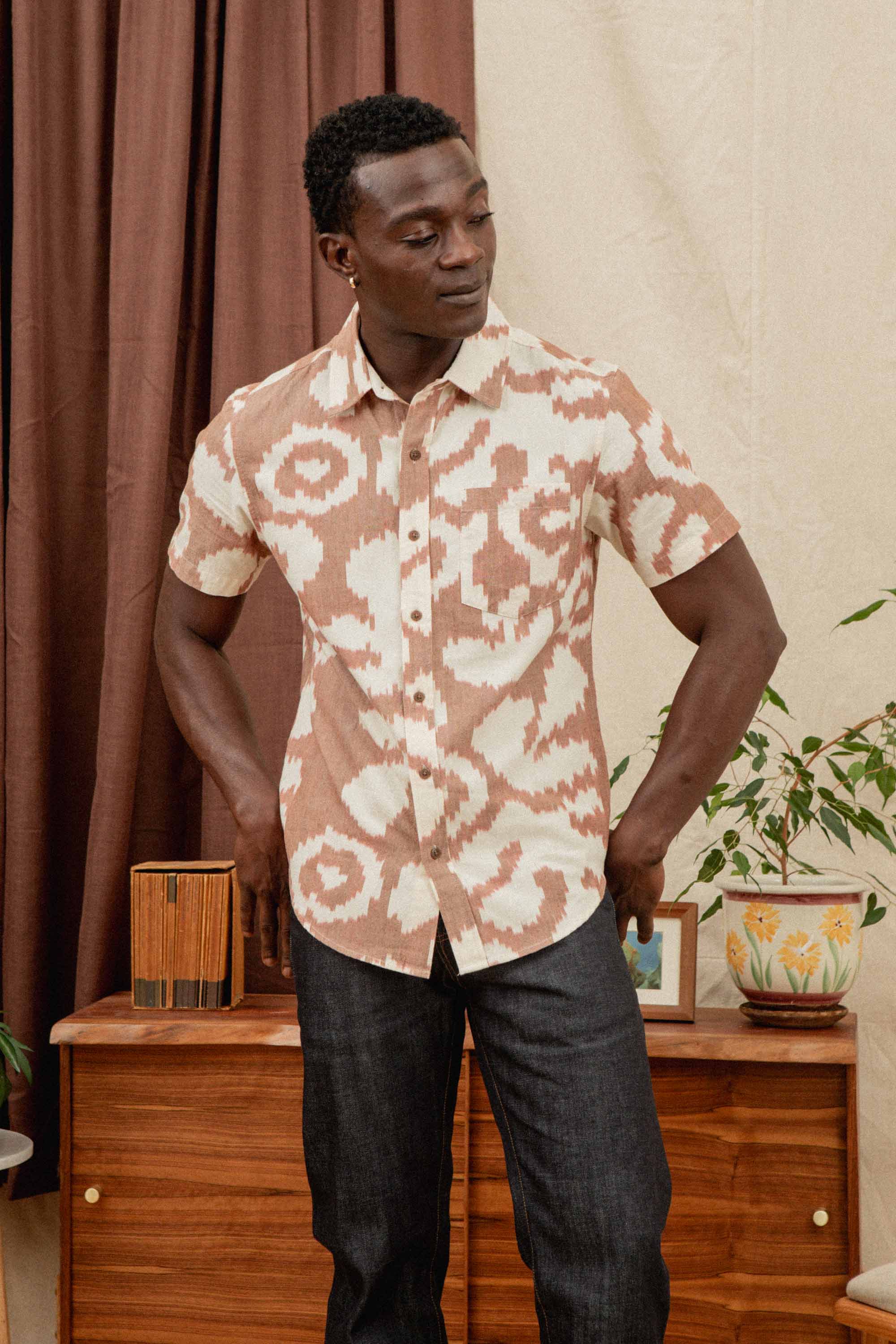 A man wears the Bridge & Burn Harbor Slim Shirt in Pop Leaf, styled with dark jeans, standing indoors before a wooden dresser decorated with plants and decor items.