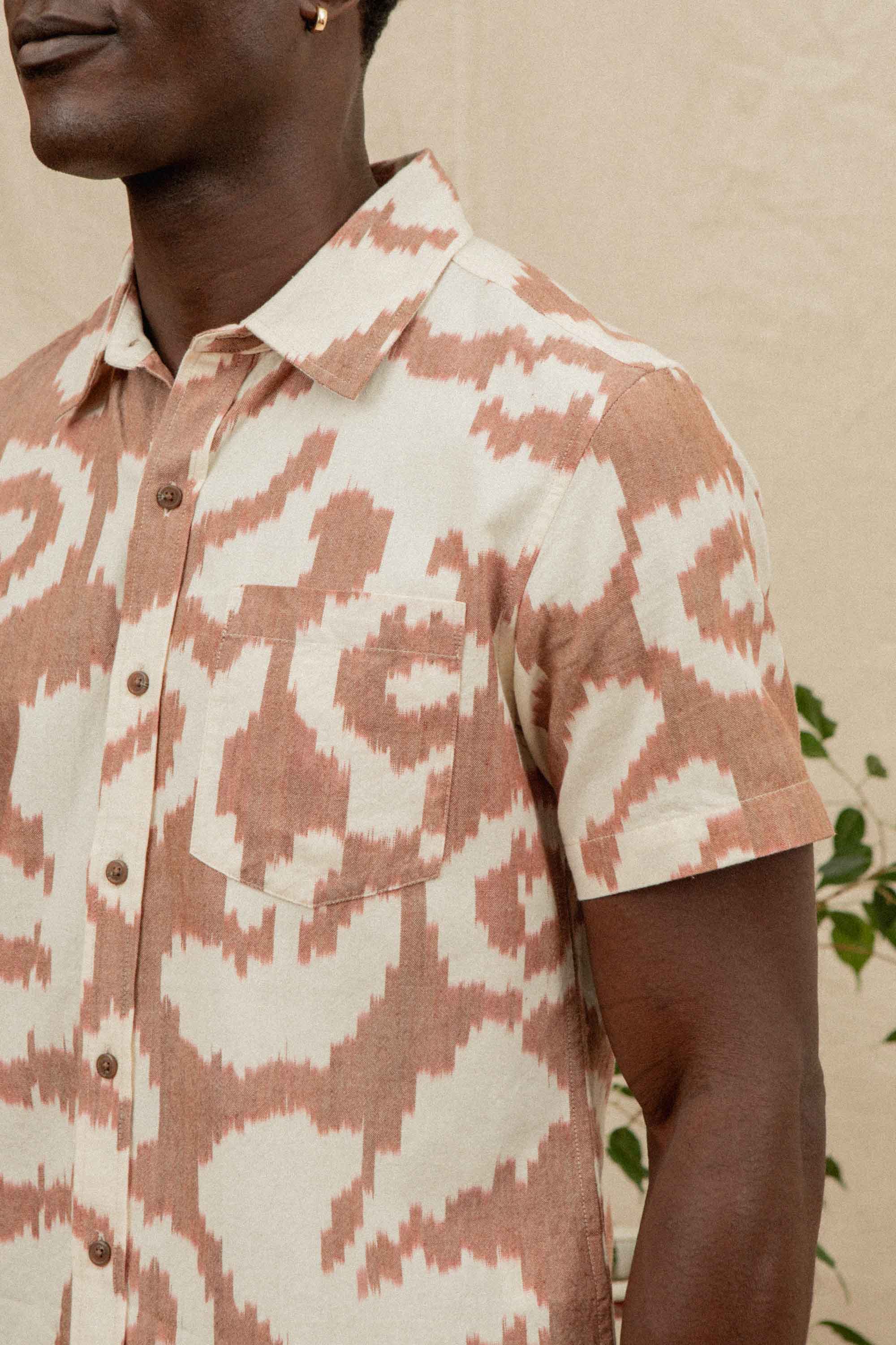 Wearing the Bridge & Burn Harbor Slim Shirt / Pop Leaf—a slim tailored, short-sleeved button-up with a beige and rust abstract pattern—a person stands before a neutral backdrop with green leafy plants.