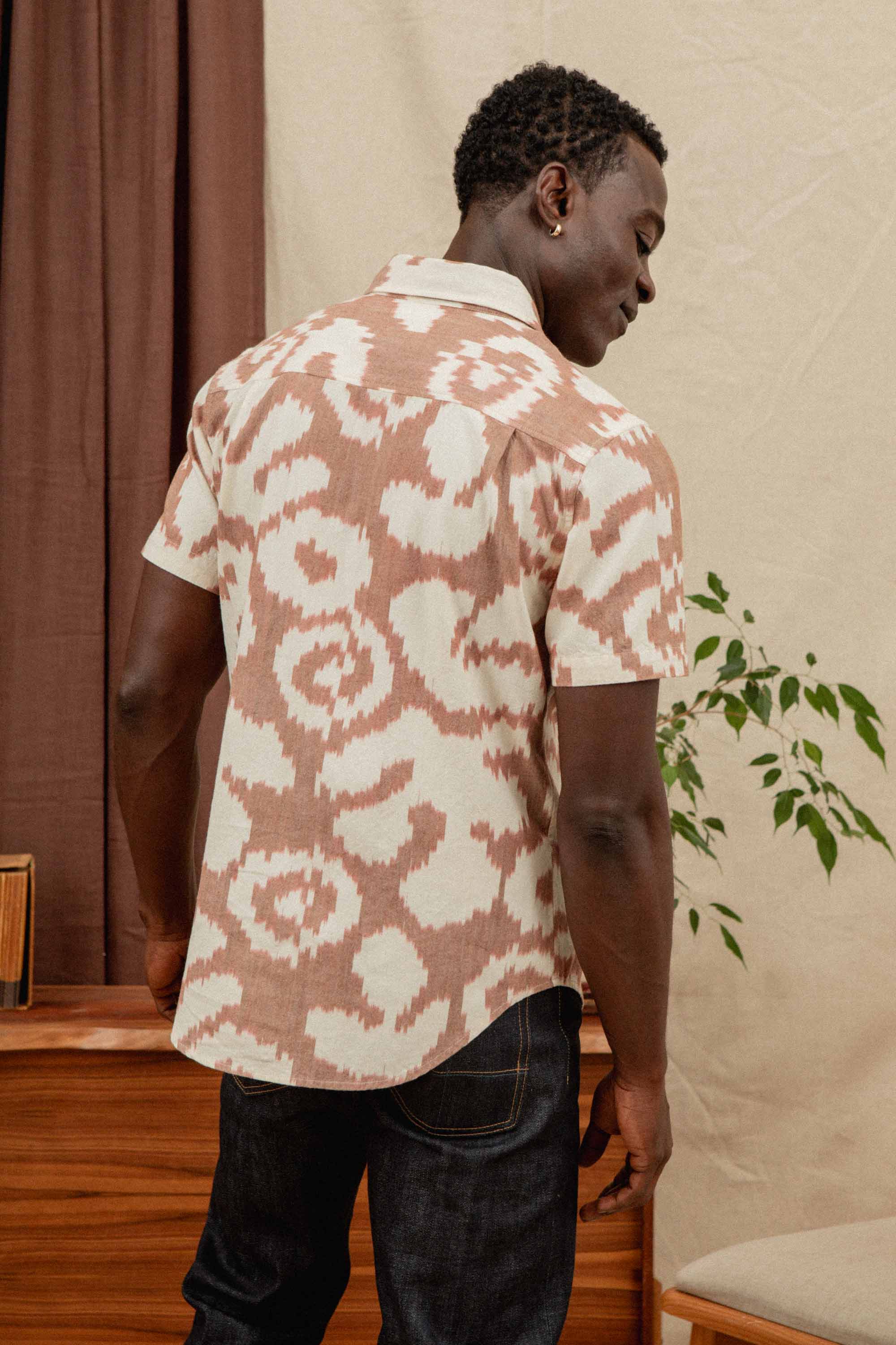 A man stands indoors with his back to the camera, wearing the Bridge & Burn Harbor Slim Shirt in Pop Leaf print and dark jeans, near a wooden table with a plant and brown curtains in the background.