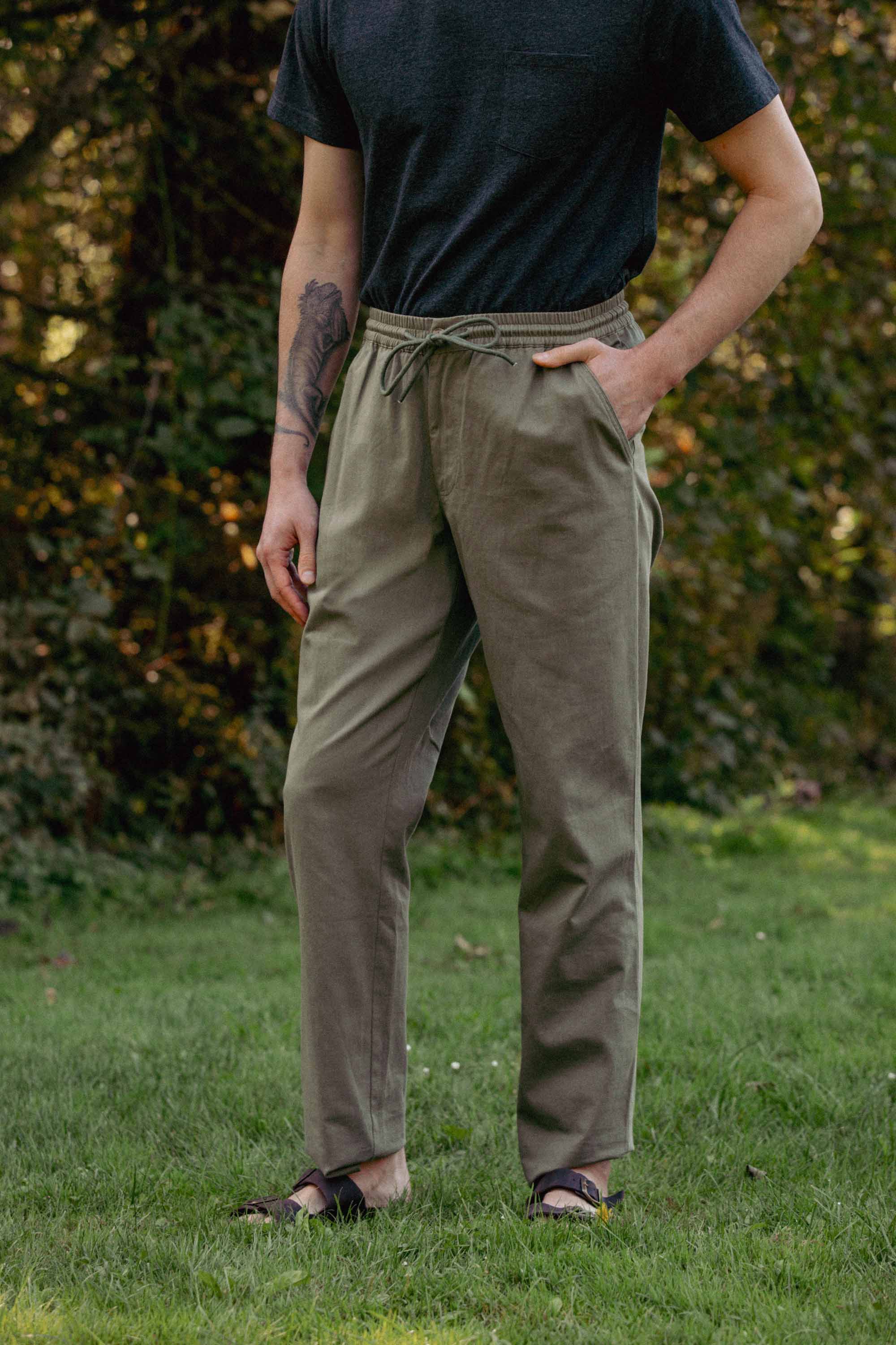 Standing outdoors on grass, a person wears Bridge & Burn’s Hayes Pant in Olive—relaxed fit with an elastic waistband—paired with a dark short-sleeve shirt. Hand in pocket, they show part of a tattoo amid a leafy, casual backdrop.