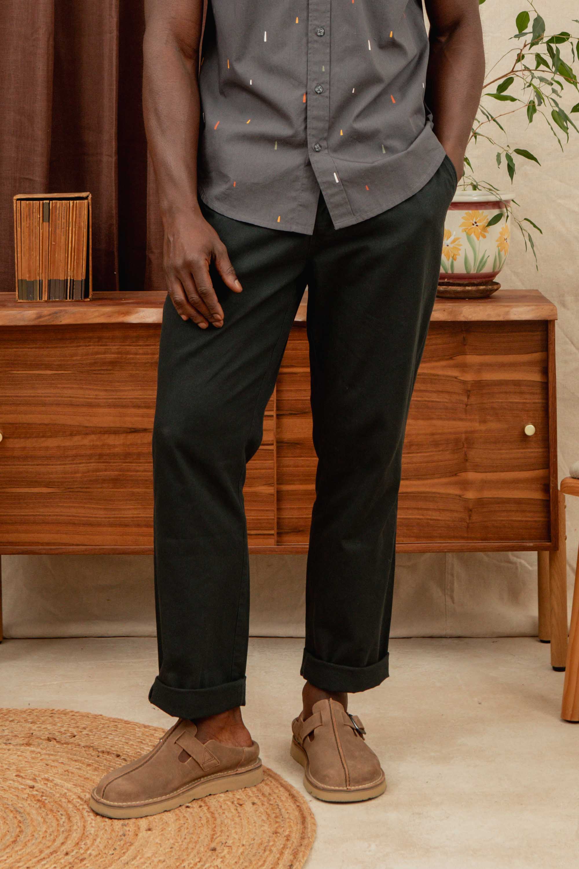 A person stands indoors wearing the Bridge & Burn Hayes Pant in Soft Black. In the background are a wooden dresser, potted plant, and woven rug. The person’s head is not visible in the frame.