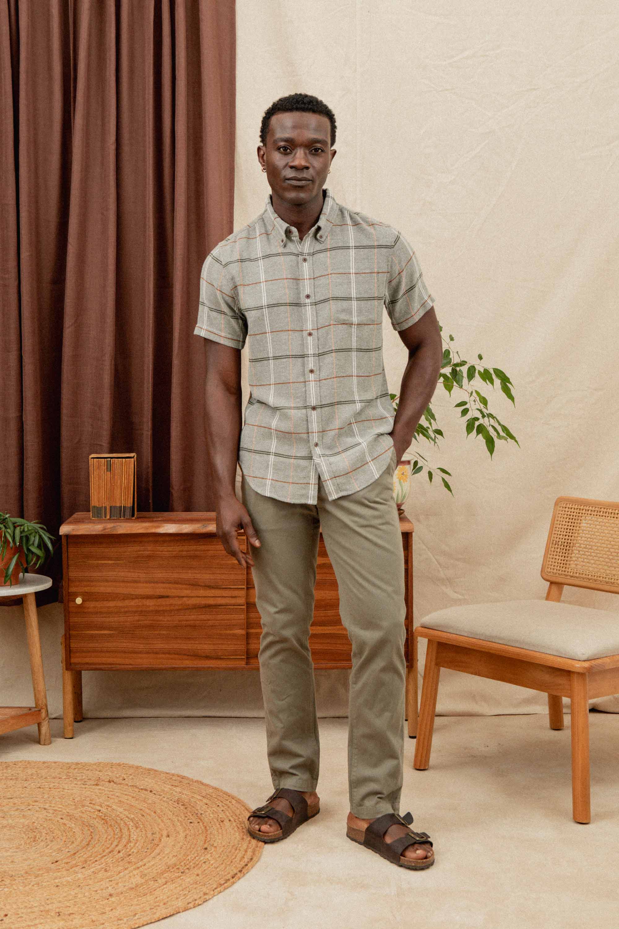 A man wears the Bridge & Burn Jordan Slim Shirt in Olive Ember Grid, paired with olive pants, posing indoors by mid-century furniture, a plant, and brown curtains above a neutral rug.