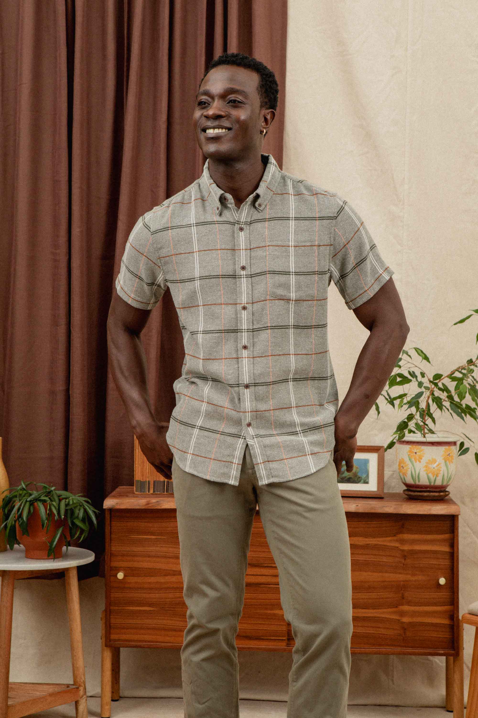 A smiling man stands indoors wearing the Bridge & Burn Jordan Slim Shirt in Olive Ember Grid and khaki pants, in front of a wooden cabinet with potted plants, a brown curtain, and a cream-colored wall.