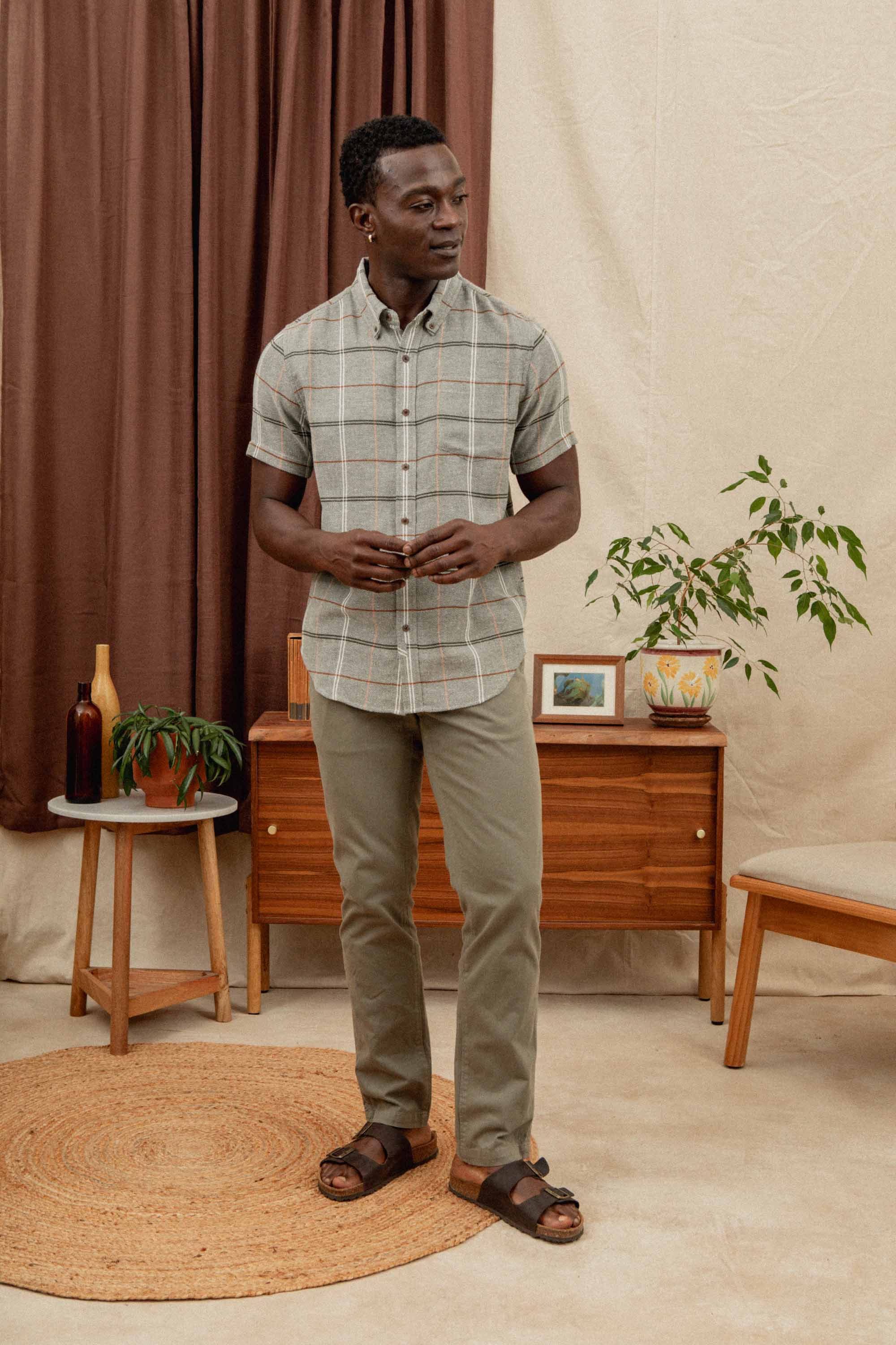 A man stands indoors wearing the Bridge & Burn Jordan Slim Shirt in Olive Ember Grid, paired with olive pants and brown sandals, in a cozy room featuring wooden furniture, potted plants, framed art, and a round woven rug.