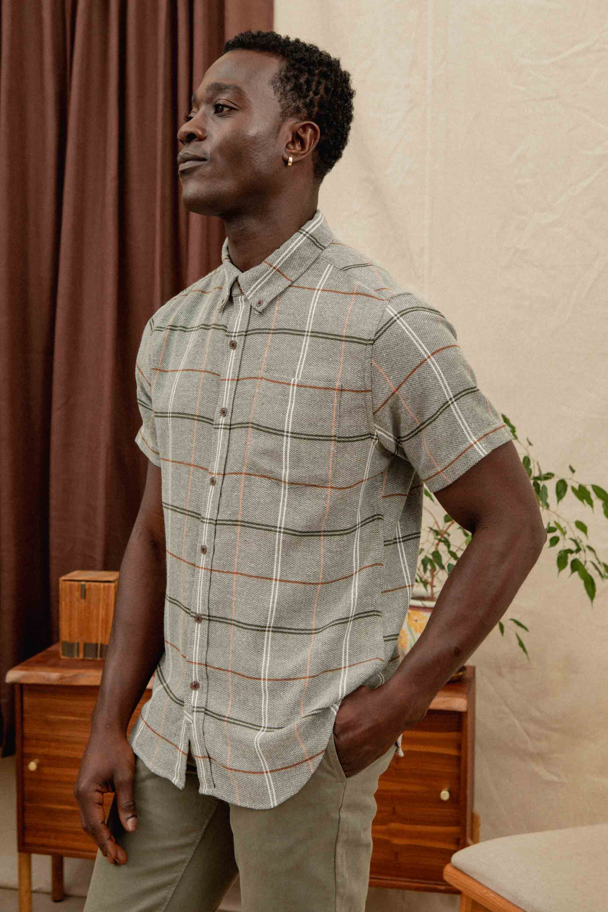 A man wearing the Bridge & Burn Jordan Slim Shirt in Olive Ember Grid stands indoors with one hand in his pocket, looking to the side. Brown curtains and a wooden table with a plant are visible in the background.