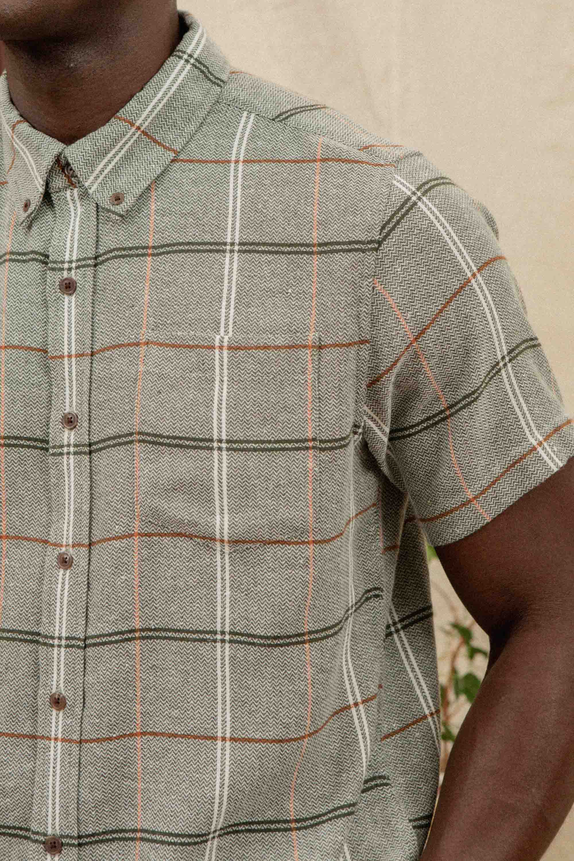 A person wears the Bridge & Burn Jordan Slim Shirt in Olive Ember Grid, featuring a green and white plaid with thin orange lines and short sleeves. The image captures the upper body from neck to mid-torso.