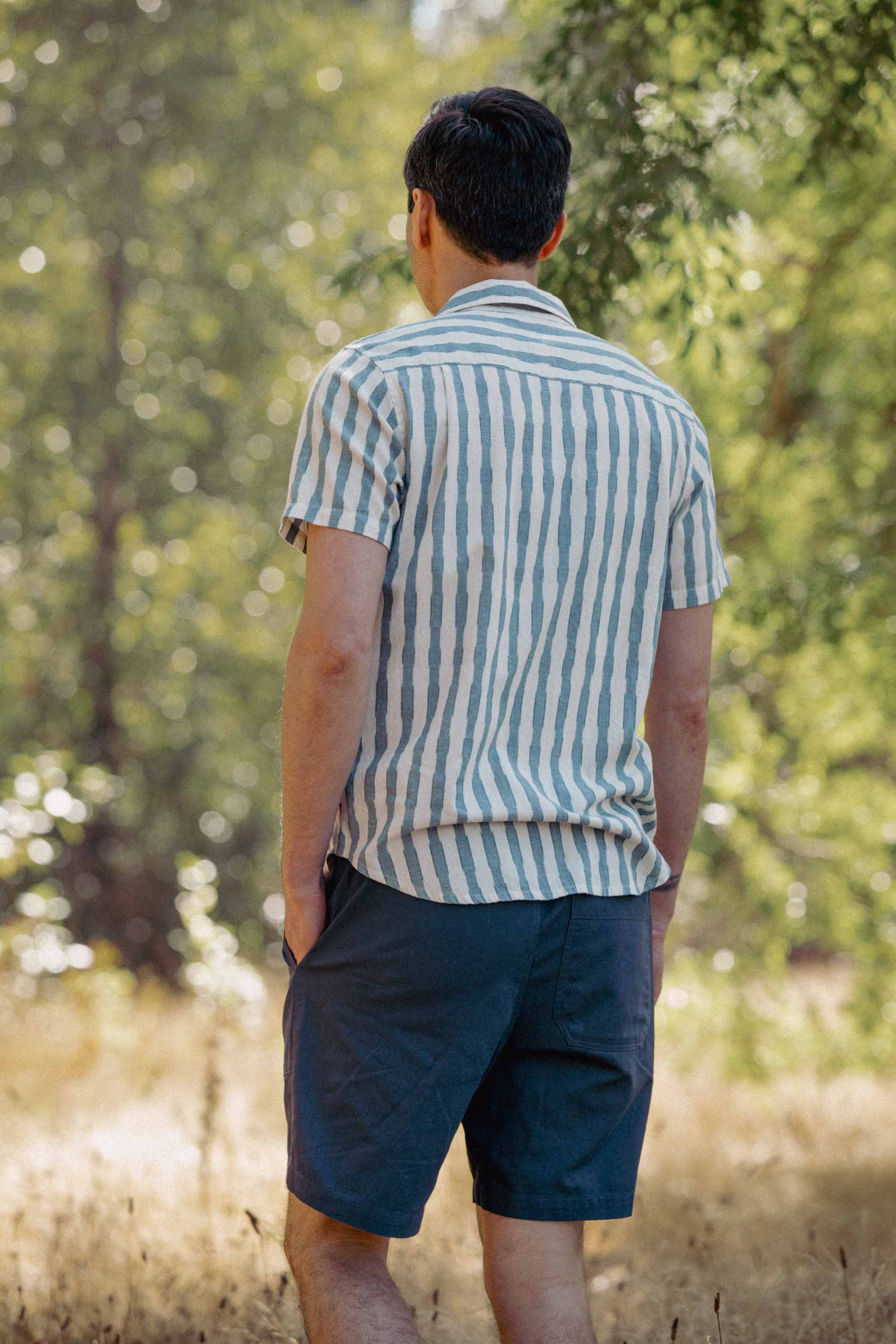 A person stands outdoors with their back to the camera, wearing the Bridge & Burn Marten Shirt in Beach Stripe and dark blue shorts. Their hands are in their pockets as trees and sunlight fill the background.