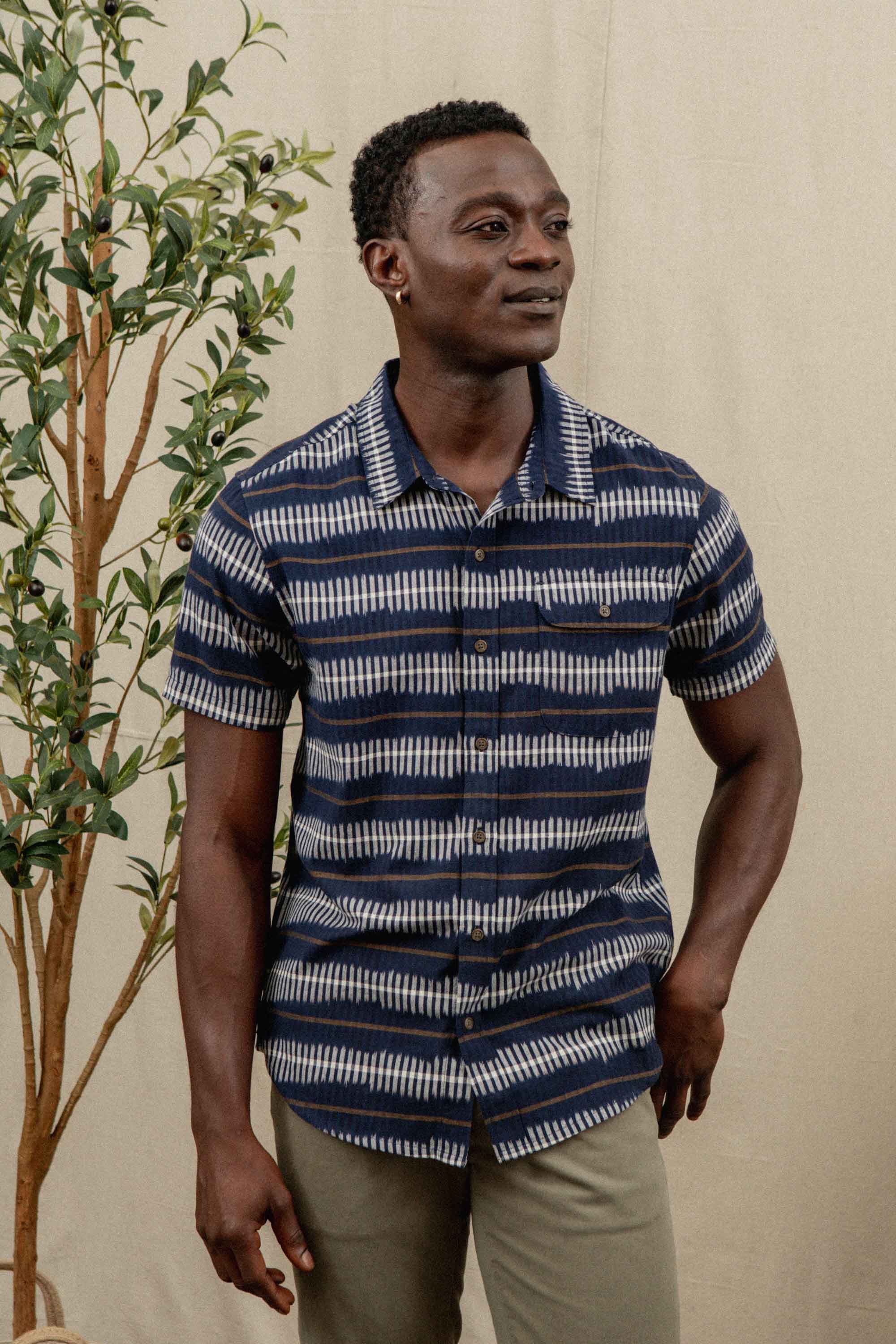 A man wearing the Bridge & Burn Marten Shirt in Dusk Mirage stands in front of a beige backdrop with a leafy plant beside him, paired with light khaki pants.