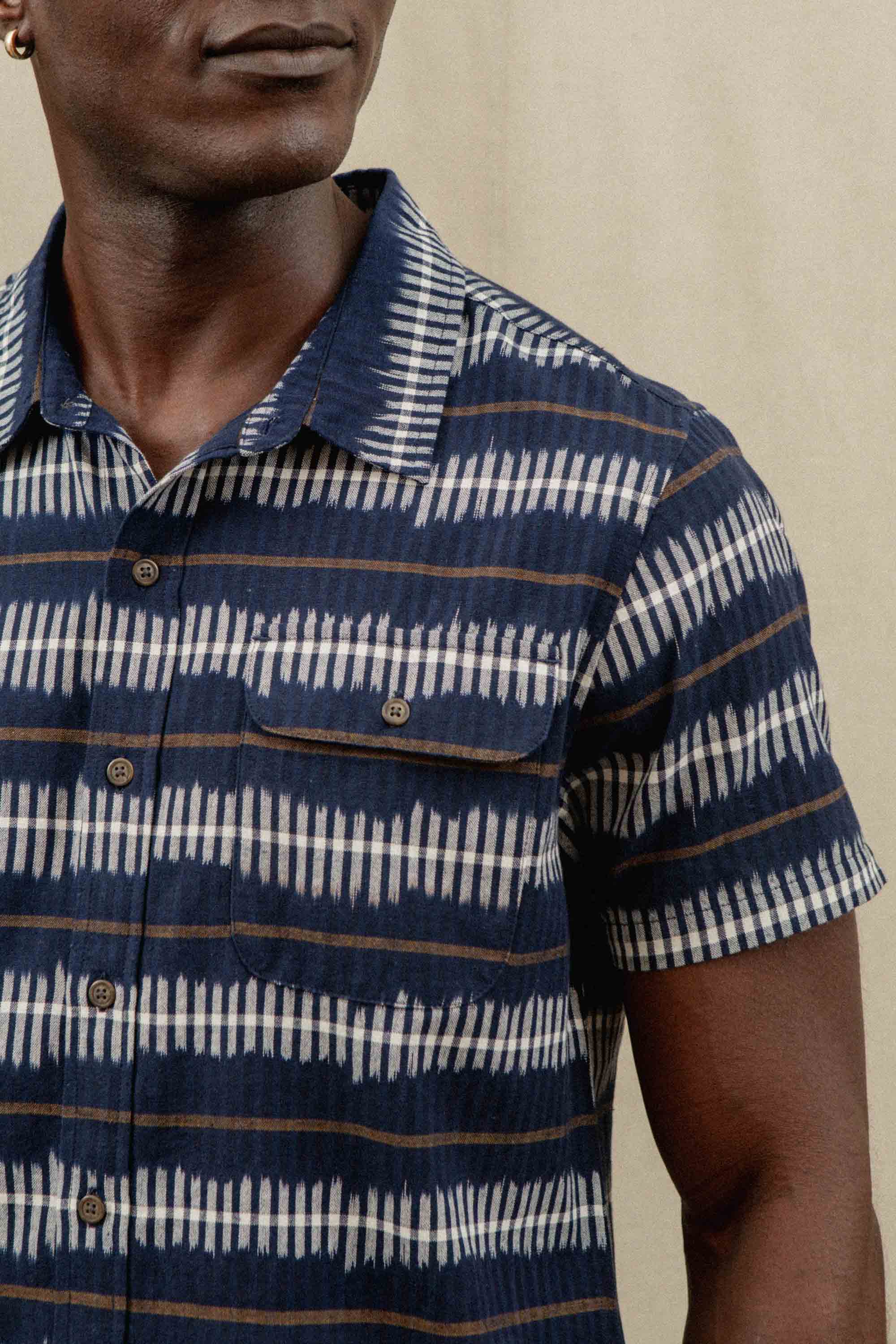 A man wears the Bridge & Burn Marten Shirt in Dusk Mirage—a classic fit, short sleeve button-down with navy, white, and brown stripes—standing against a neutral background. Only his upper body and part of his face are visible.