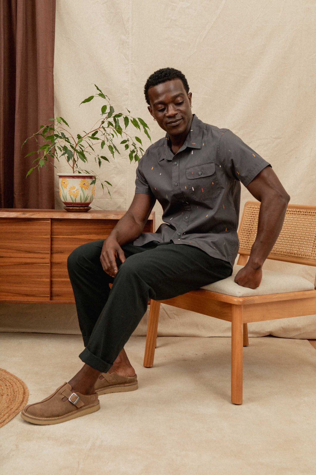 A man with a limb difference, wearing the Bridge & Burn Marten Shirt in Midnight Rain, black pants, and brown shoes, sits on a wooden chair beside a mid-century cabinet with a potted plant. He smiles slightly while looking down.