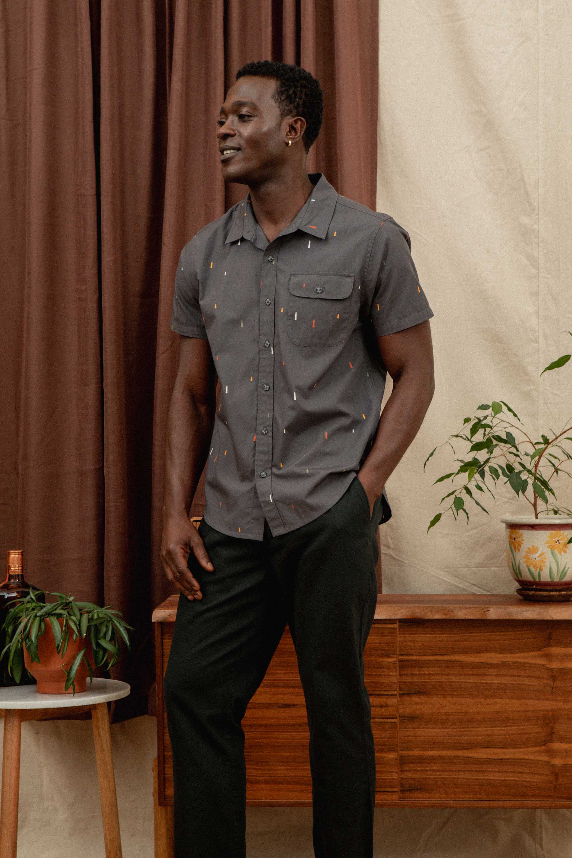 A man stands indoors smiling with one hand in his pocket, wearing the Bridge & Burn Marten Shirt in Midnight Rain and dark pants. Behind him are brown curtains, a wooden console, and potted plants.