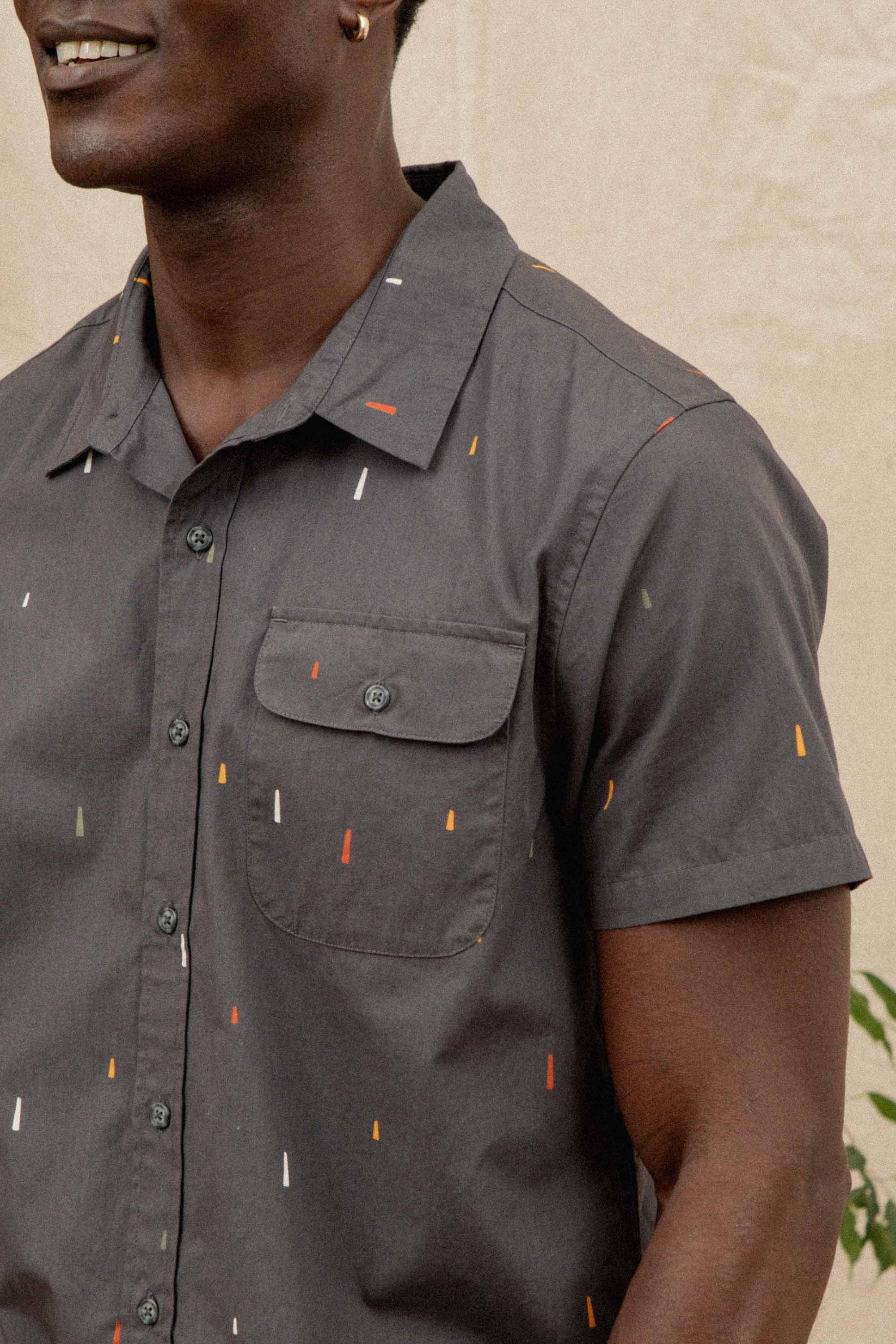 A person wears the Bridge & Burn Marten Shirt in Midnight Rain—a classic fit, short sleeve button-down in gray with a chest pocket and a subtle small, colorful dash pattern. Only their lower face and upper body are visible.