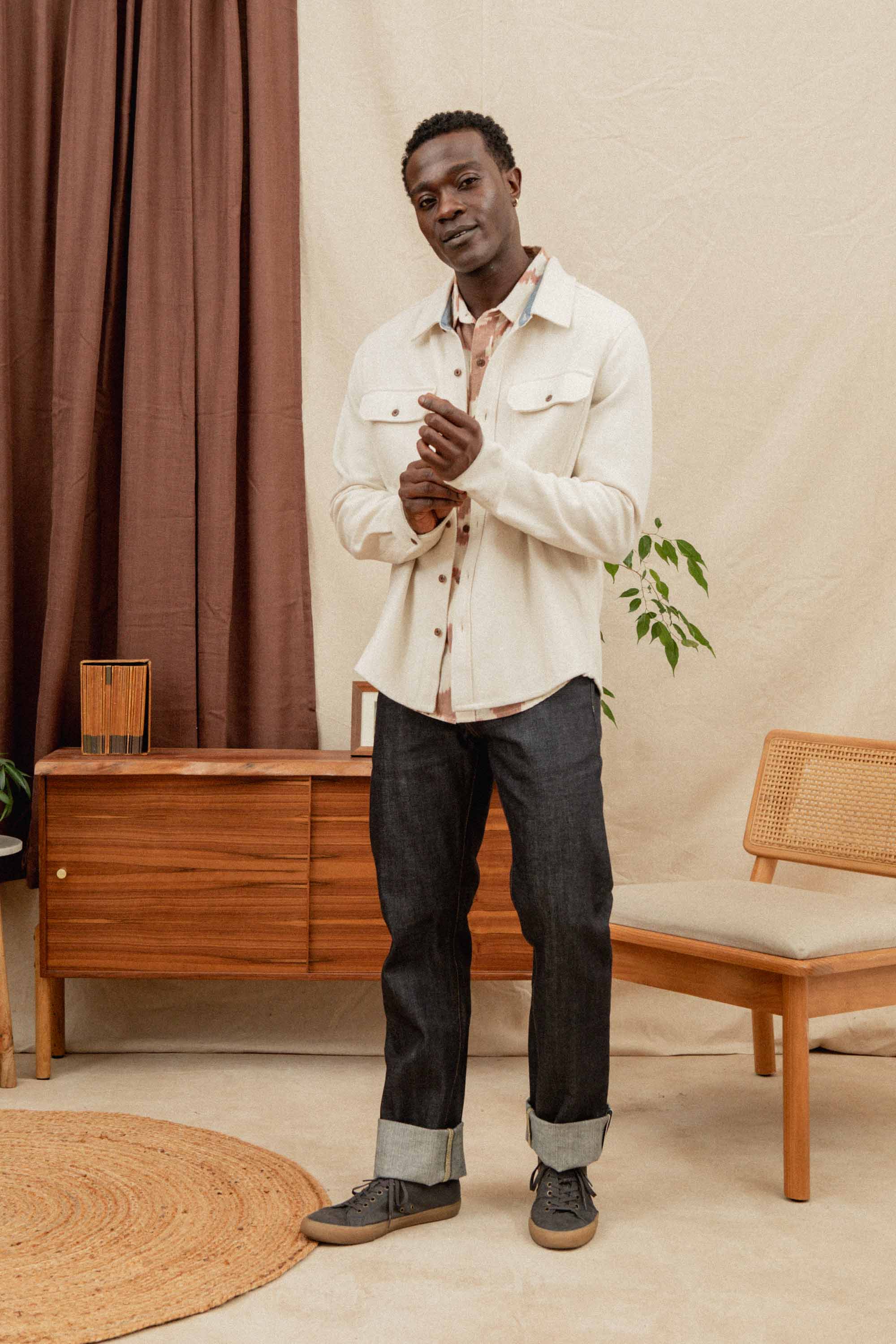 A man stands indoors on a circular rug, wearing the Bridge & Burn Pointer Overshirt in Ivory Twill over a tan shirt, dark cuffed jeans, and gray sneakers. Behind him are a wooden sideboard, chair, potted plant, and brown curtains.