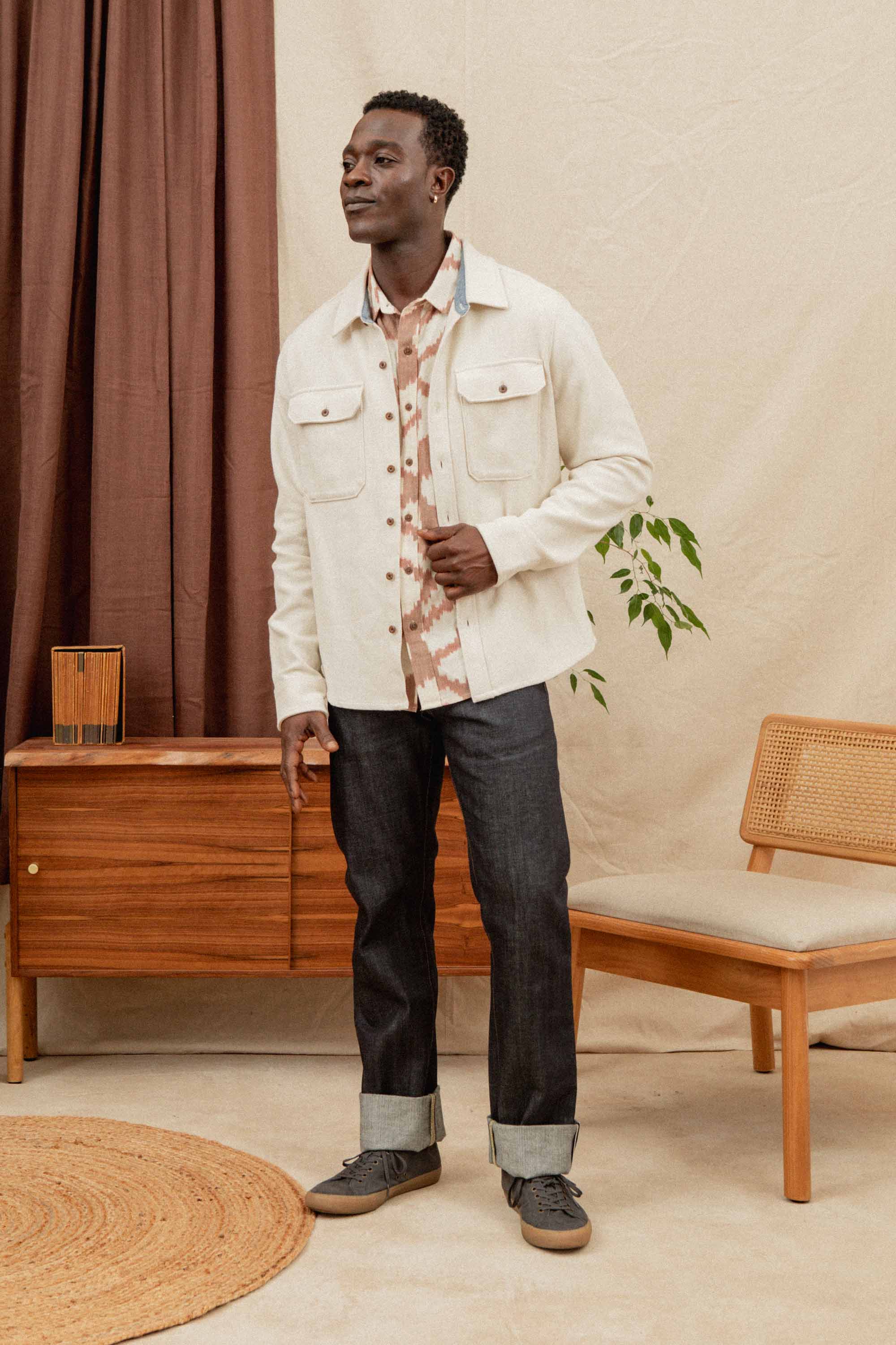 A man wears the Bridge & Burn Pointer Overshirt in Ivory Twill over a patterned shirt, paired with dark cuffed jeans and gray sneakers. He smiles indoors, holding his jacket, with a wooden cabinet, chair, and potted plant behind him.