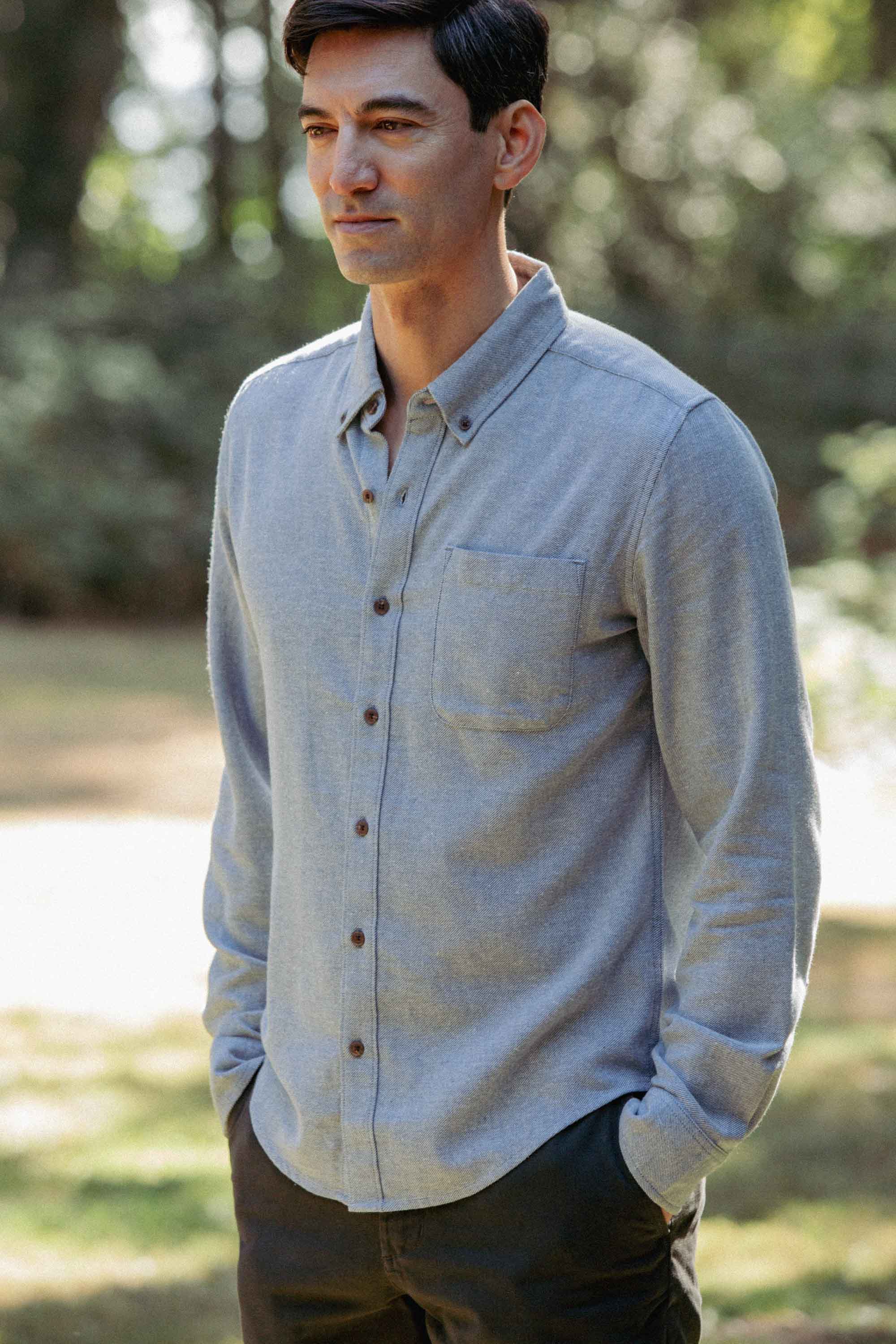 A man stands outdoors in a sunlit, wooded area wearing the Bridge & Burn Sutton Slim Shirt in Denim Twill with dark pants. He has short dark hair and his hands are in his pockets.