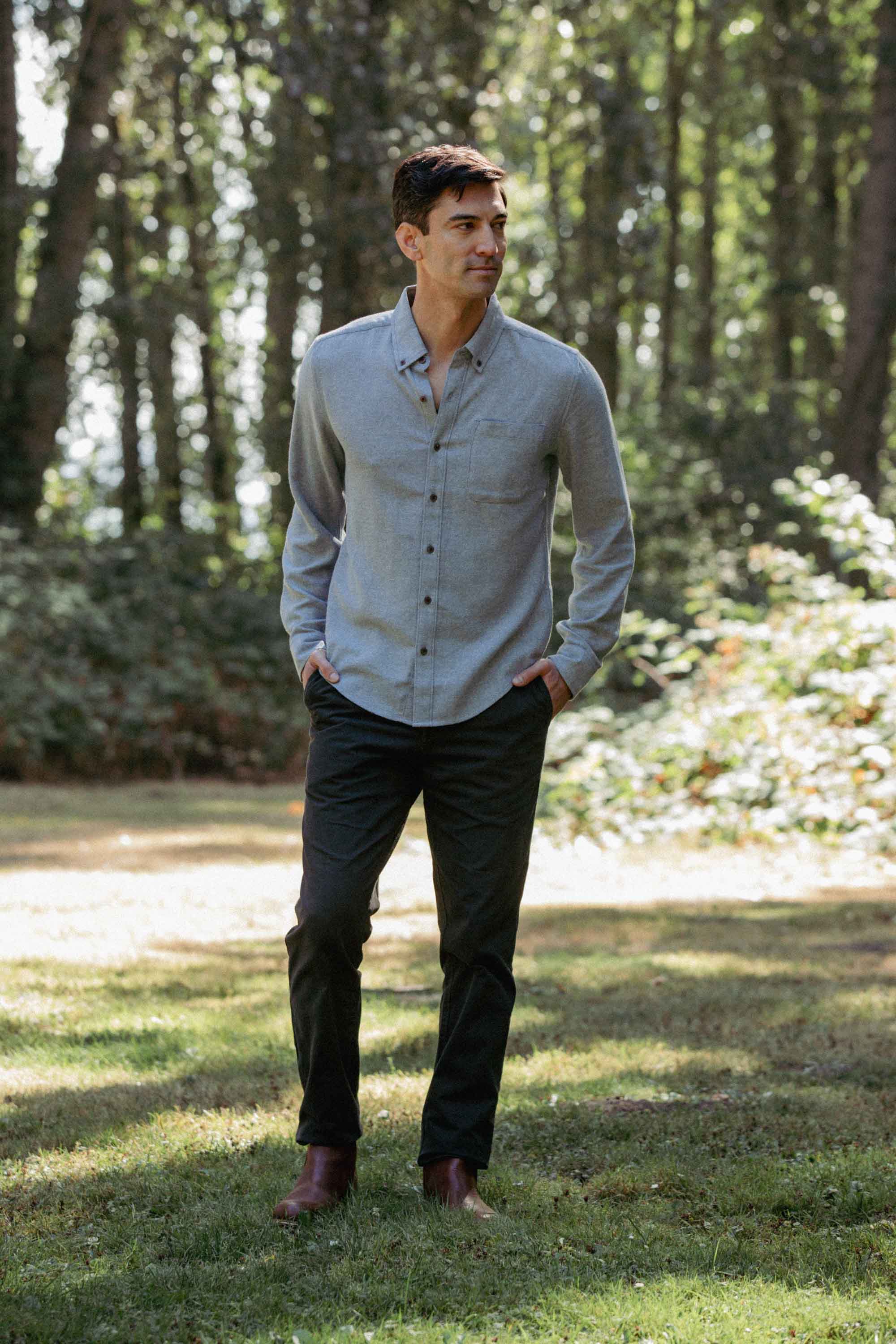 A man stands on grass, wearing the Bridge & Burn Sutton Slim Shirt in Denim Twill with dark pants and brown shoes. Sunlight filters through tall trees in the background as he puts his hands in his pockets.