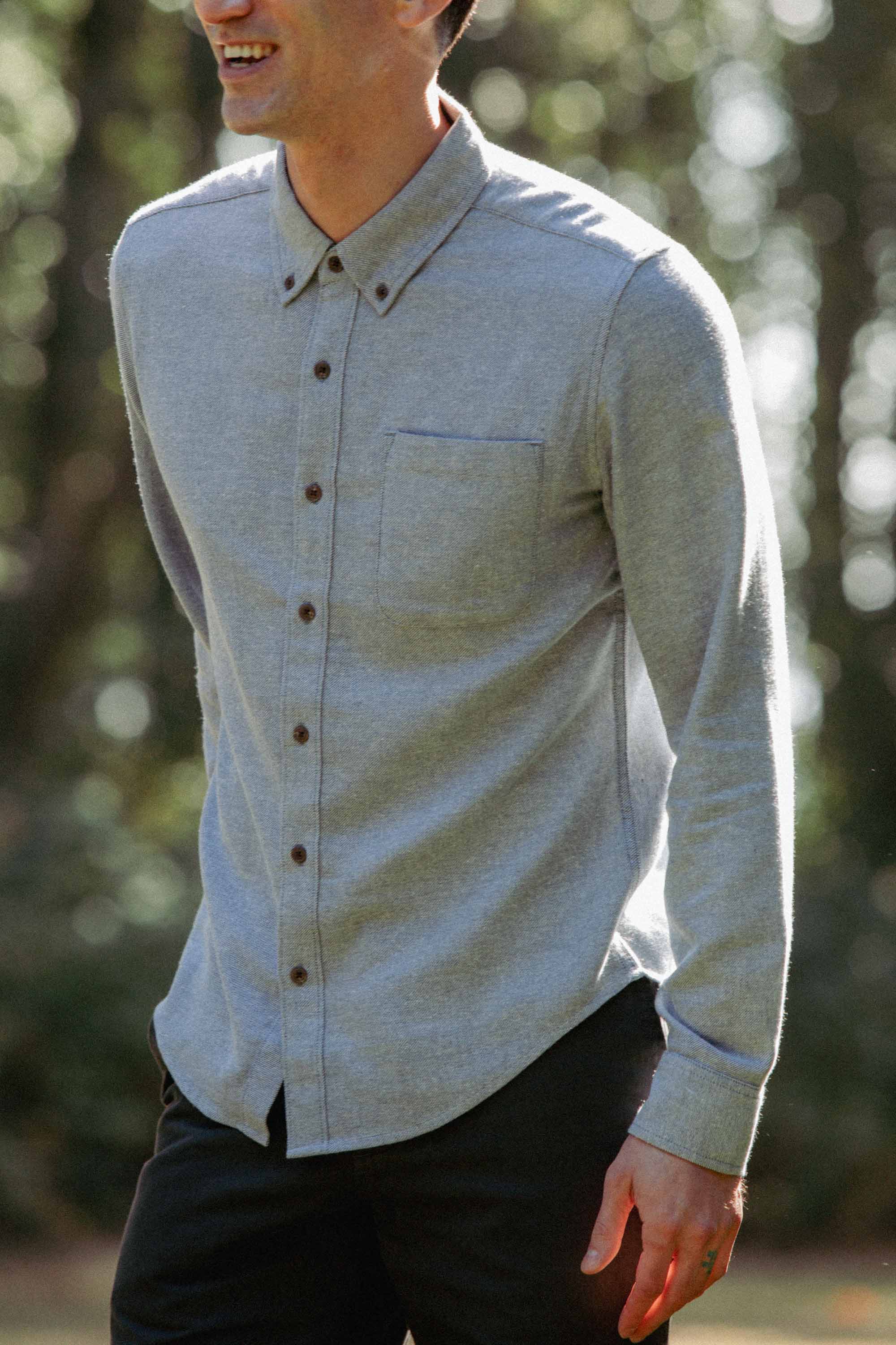 A person wears the Bridge & Burn Sutton Slim Shirt in Denim Twill with black pants, standing outdoors and smiling amid blurred greenery. The image is cropped above the chin, showing the shirt's upper body style.