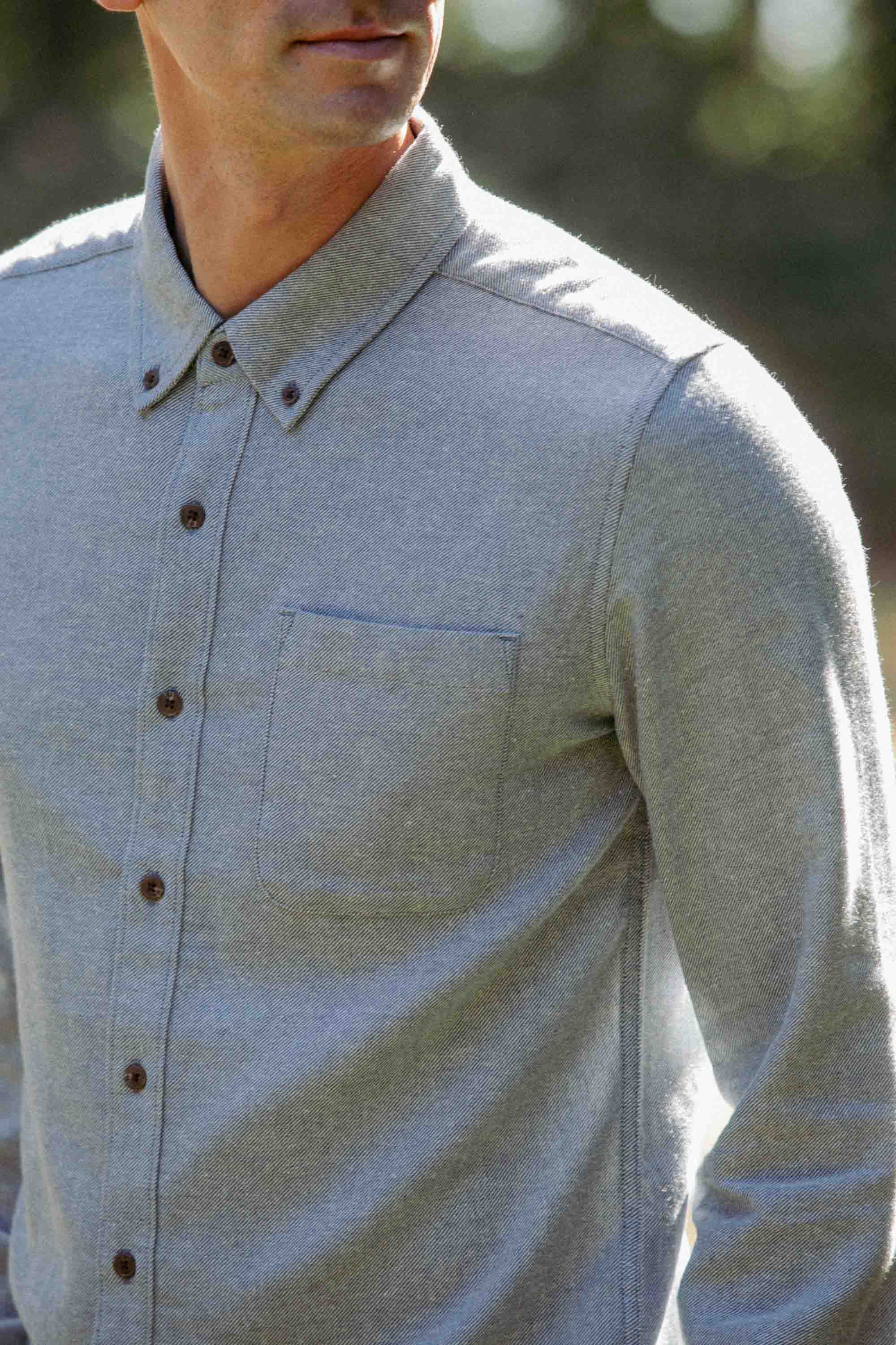 A man wearing the Bridge & Burn Sutton Slim Shirt in Denim Twill is standing outdoors with blurred greenery in the background, his face partially visible as he turns slightly to the side.